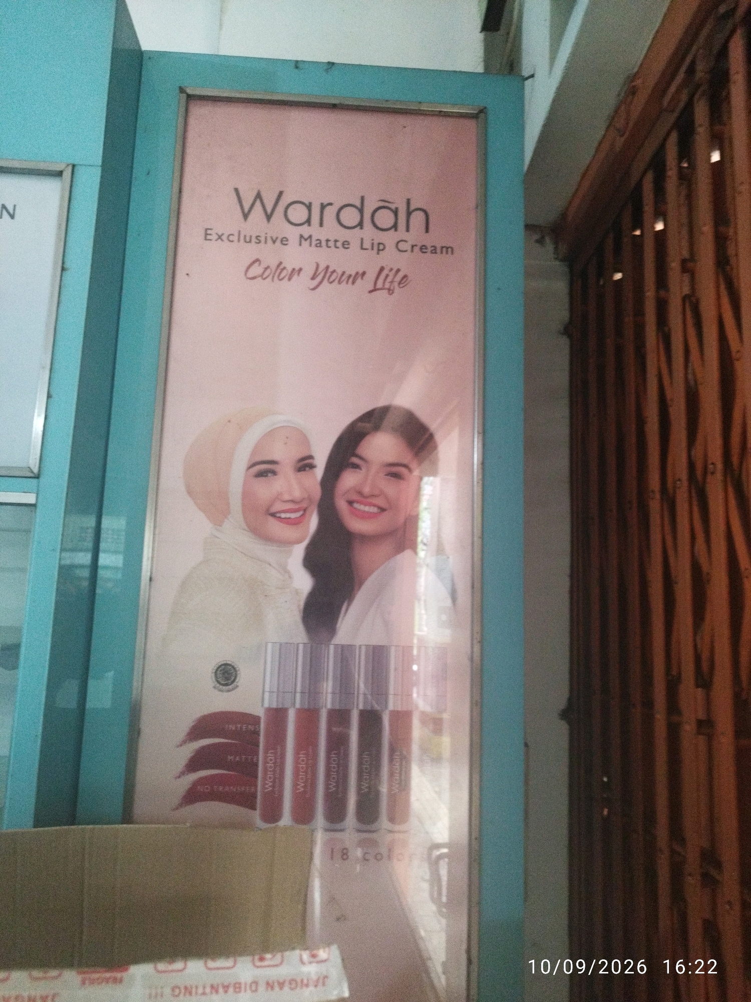 The image shows a vertical banner advertisement for Wardah Exclusive Matte Lip Cream, featuring two smiling women. The banner is framed by a light blue border and is set against a pink background with text. Below the women, there are four lipstick tubes and the text "18 colors". A cardboard box is partially visible in the lower left corner, with "FRAGILE" and Indonesian text printed on a white tape. The timestamp "10/09/2026 16:22" is visible in the lower right corner. The overall setting appears to be outdoors, possibly on a street in Mojokerto, Indonesia, with a corrugated metal structure in the background on the right.