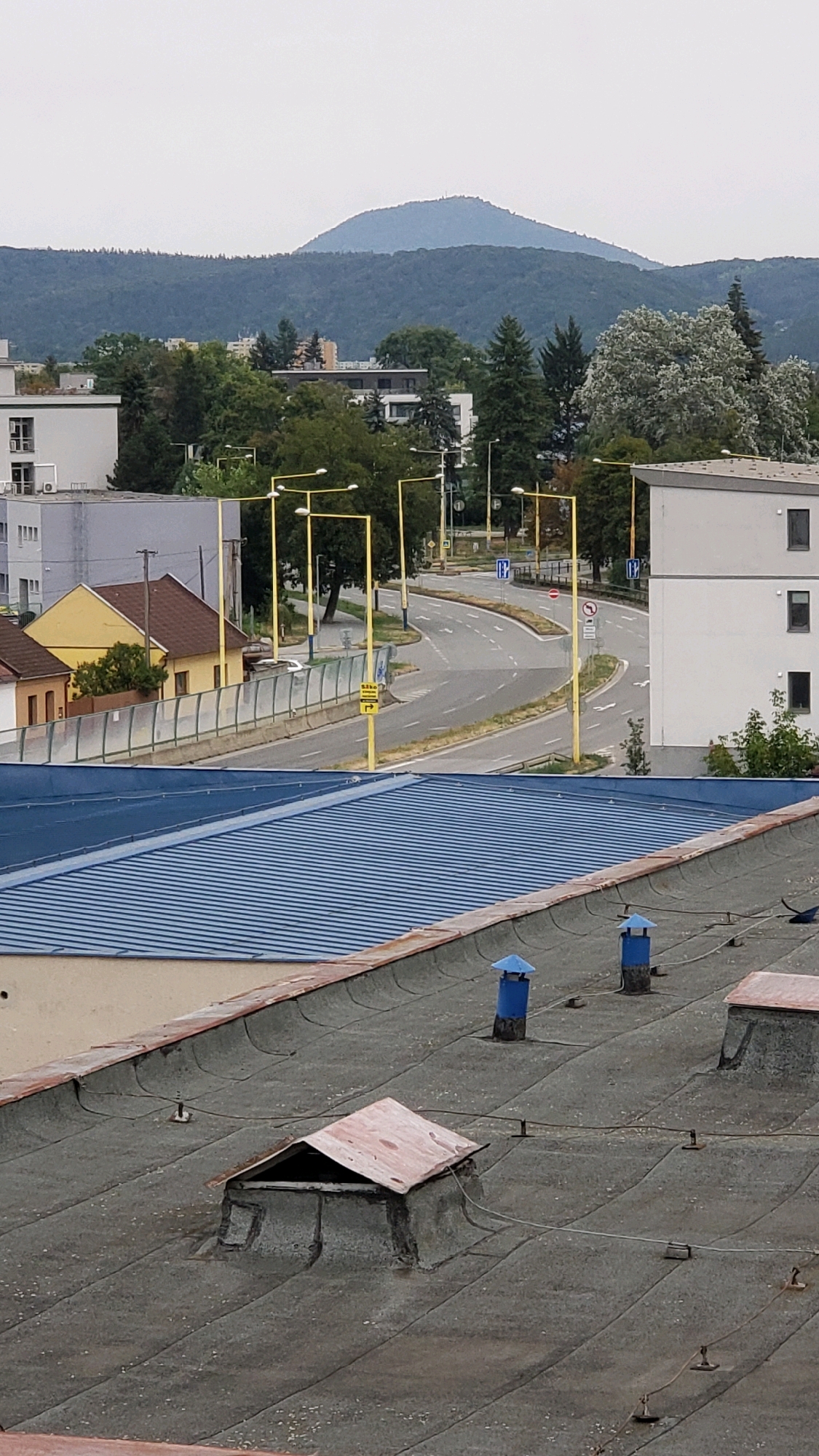 The image captures a split perspective of an urban environment. The upper portion displays a broad view of Prešov, Slovakia, featuring a prominent, hazy blue mountain range in the distance, likely Zemplínske vrchy. In the midground, a winding road with multiple lanes is visible, bordered by yellow streetlights and various buildings, including a bright yellow house. Trees and smaller structures populate the landscape between the road and the mountains. The lower section of the image shifts focus to the rooftops of buildings. A dark, weathered flat roof occupies the foreground, adorned with several vent pipes, some with blue caps, and a distinctive, dilapidated pitched skylight with a red roof. A corrugated blue metal roof of another building is partially visible in the upper portion of this section. The scene is overcast, suggesting a cloudy day. There are no visible people or animals in the frame.