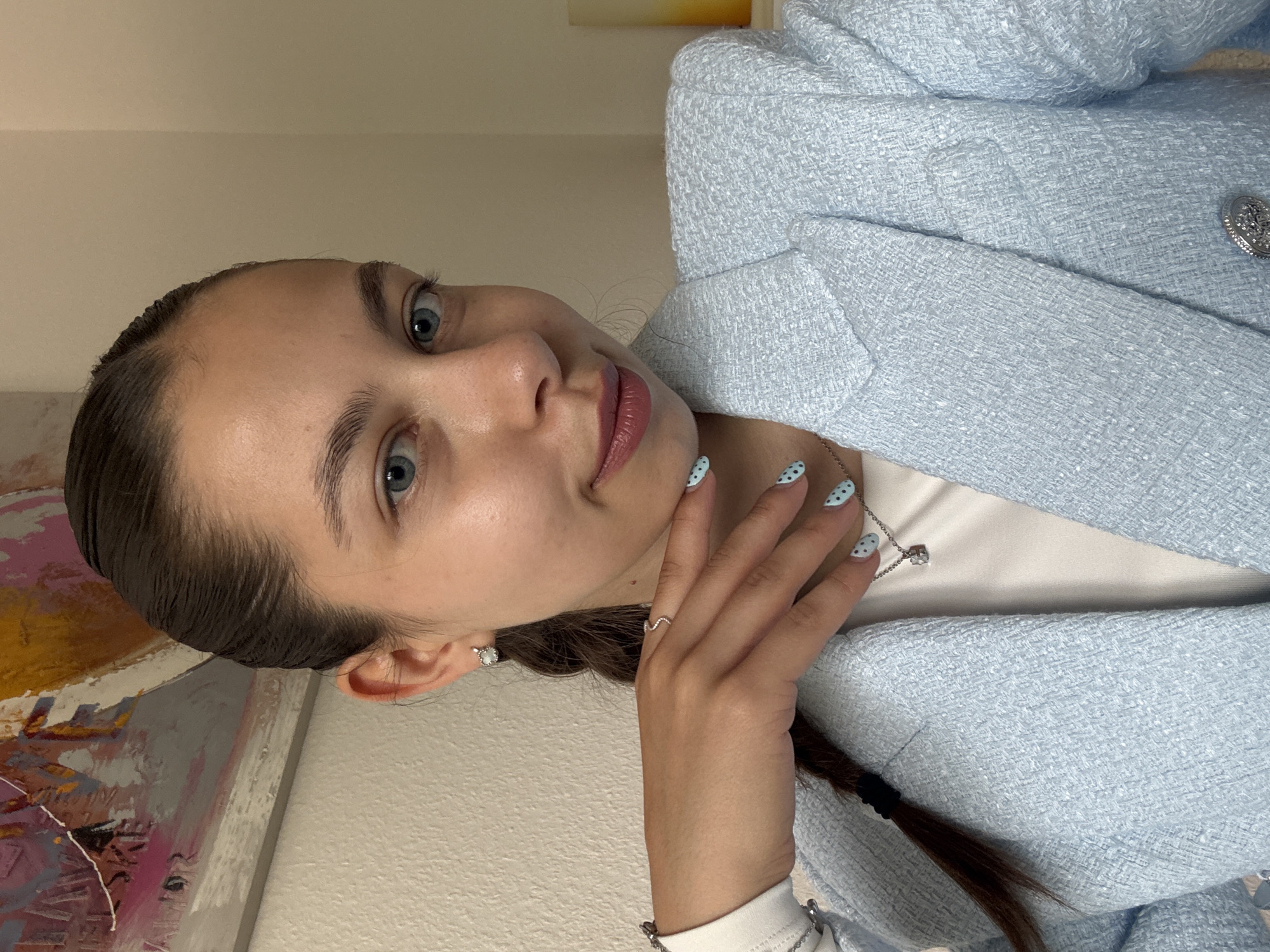 A woman is depicted in a close-up, tilted view. She has light skin, blue eyes, and her dark hair is pulled back into a braid. She wears a light blue tweed blazer over a white top, and a necklace with a pendant is visible. Her nails are painted light blue with black polka dots. She is wearing earrings and a ring. The background includes a partially visible painting and a light-colored wall. There is no information to place this image in Bruchköbel, Germany.