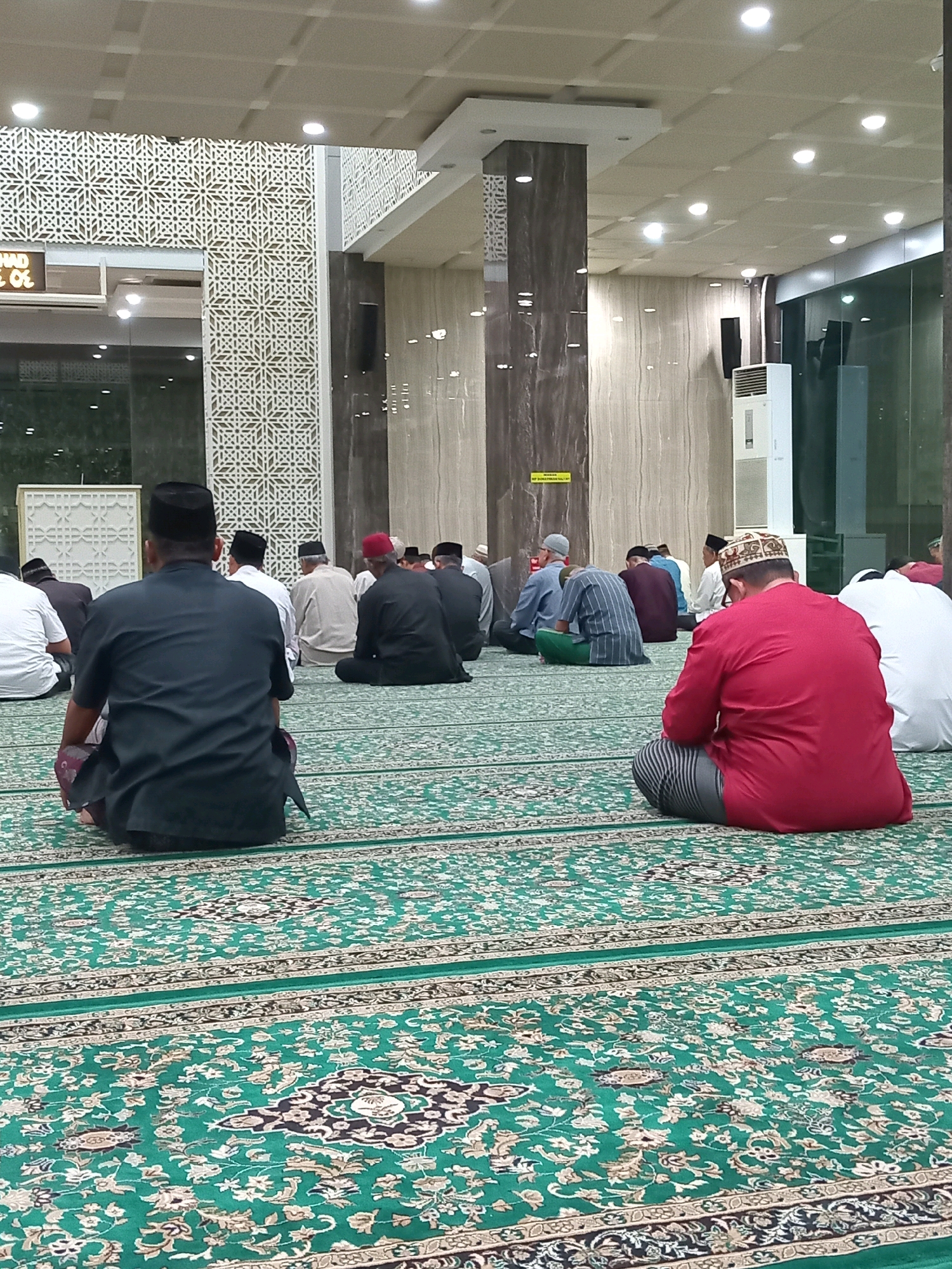 The image depicts the interior of a mosque in Randuagung, Indonesia, with several men seated on a green carpet adorned with intricate patterns. The men are facing away from the camera, suggesting they are engaged in prayer or a religious gathering. The mosque features modern architecture, with a high ceiling and large columns. A digital clock displaying "07.46" is visible in the upper left corner. The overall atmosphere appears to be one of solemnity and devotion.