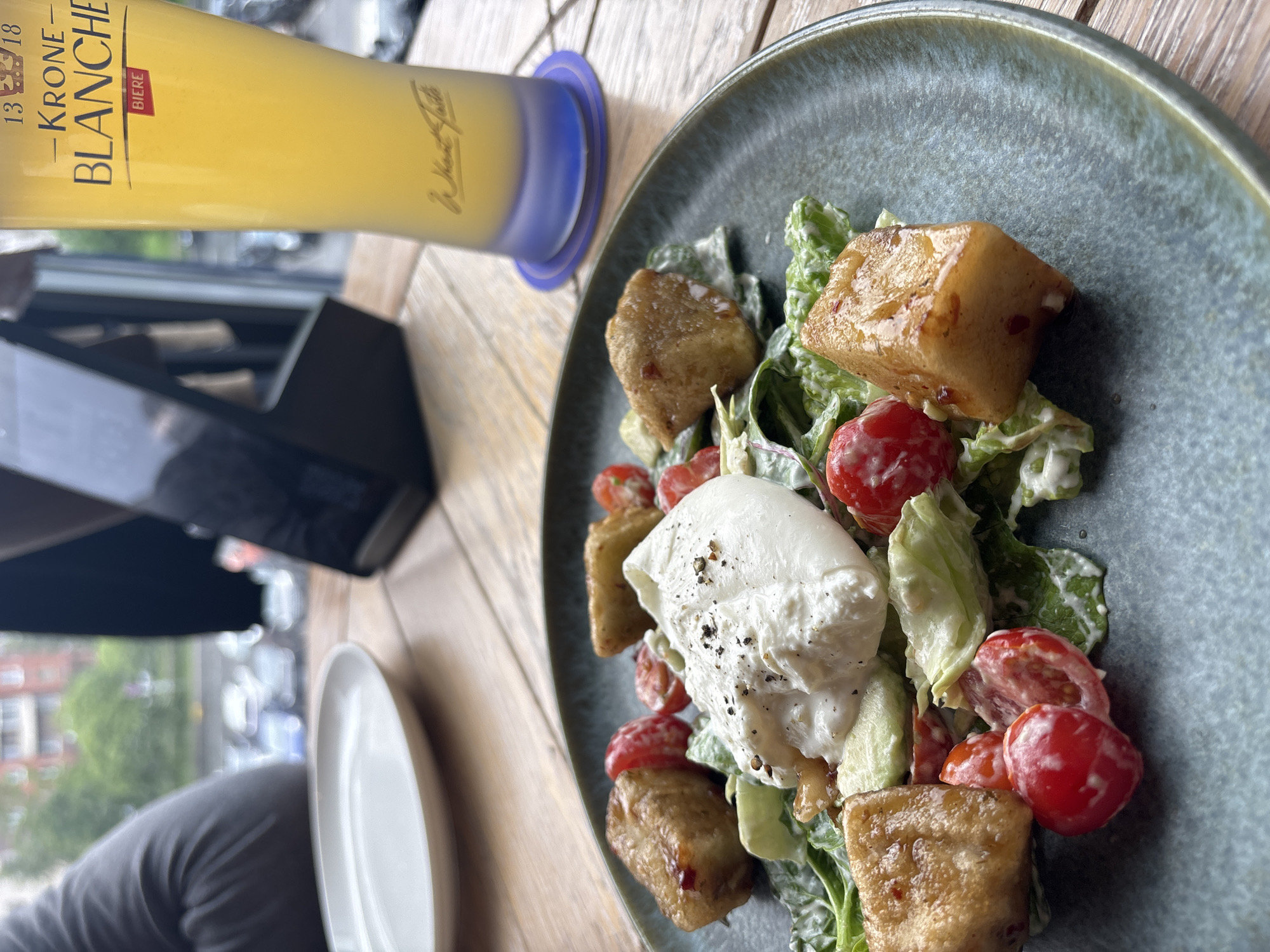The image shows a plate of food and a glass of beer on a wooden table. The plate contains a salad with tomatoes, lettuce, fried tofu, and a scoop of white cheese, possibly burrata, sprinkled with black pepper. The glass contains a light-colored beer, labelled "KRONE BLANCHE BIERE," with a blue base. A portion of a white plate is visible to the left. There's also an object which appears to be a menu and a portion of a person's head in the bottom left. The image is taken indoors, likely in a restaurant or bar. Through the window, a cityscape can be seen, featuring buildings and trees, suggesting an urban setting. The city is specified as Saint Petersburg, Russia.