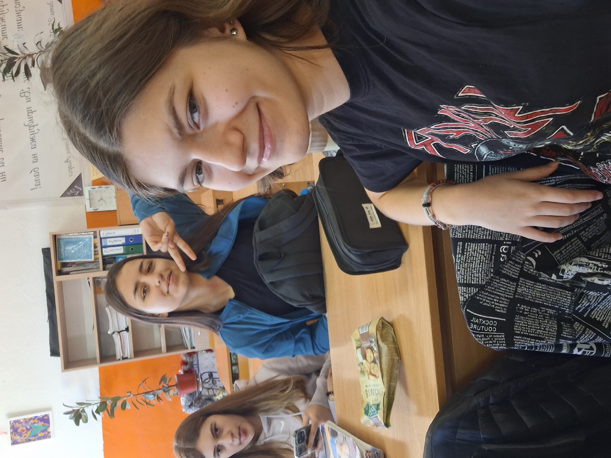 The image captures three young women in what appears to be a classroom or study setting in Panagyurishte, Bulgaria, during daytime. The environment features wooden desks, shelves filled with binders and books, and walls painted white and orange, decorated with notes and a framed picture.
In the foreground, a brunette woman with shoulder-length hair and an earring smiles directly at the camera, likely taking a selfie. She wears a black t-shirt adorned with a distinctive red and white graphic design. Her left arm rests on a black bag or patterned fabric resembling newspaper print, which displays English words like "COUTURE" and "COCKTAIL."
Behind her, another brunette woman, wearing a blue jacket, offers a peace sign and a gentle smile. A third brunette woman is visible in the background, holding a smartphone and also looking towards the camera.
On the wooden desk, a packet of snacks, possibly biscuits, with Cyrillic text including "ВЛАСГУЧ" is placed next to a black bag. A small potted plant adds a touch of nature to the scene. The overall mood is casual and friendly, suggesting a relaxed moment among classmates or friends.
Visible text on the wall includes Cyrillic script with phrases such as "дома: 5" and "мандажа на," consistent with the Bulgarian location.