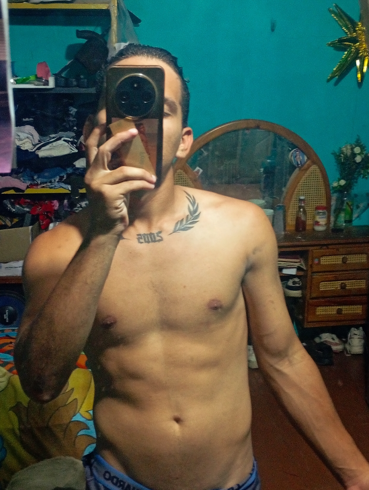 A shirtless male is shown in a medium shot, holding a smartphone in front of his face, obscuring his facial features. He appears to be taking a picture of himself. He has a tattoo of the year "2005" and a leaf design on his upper left chest. The background includes a shelf full of miscellaneous items, a mirror, a dresser with drawers, and a blue wall. A decorative star and a vase of flowers are also visible on top of the dresser. Based on the details of the setting, it is likely that the image was taken indoors in a residential environment in the city of Altagracia, Venezuela.