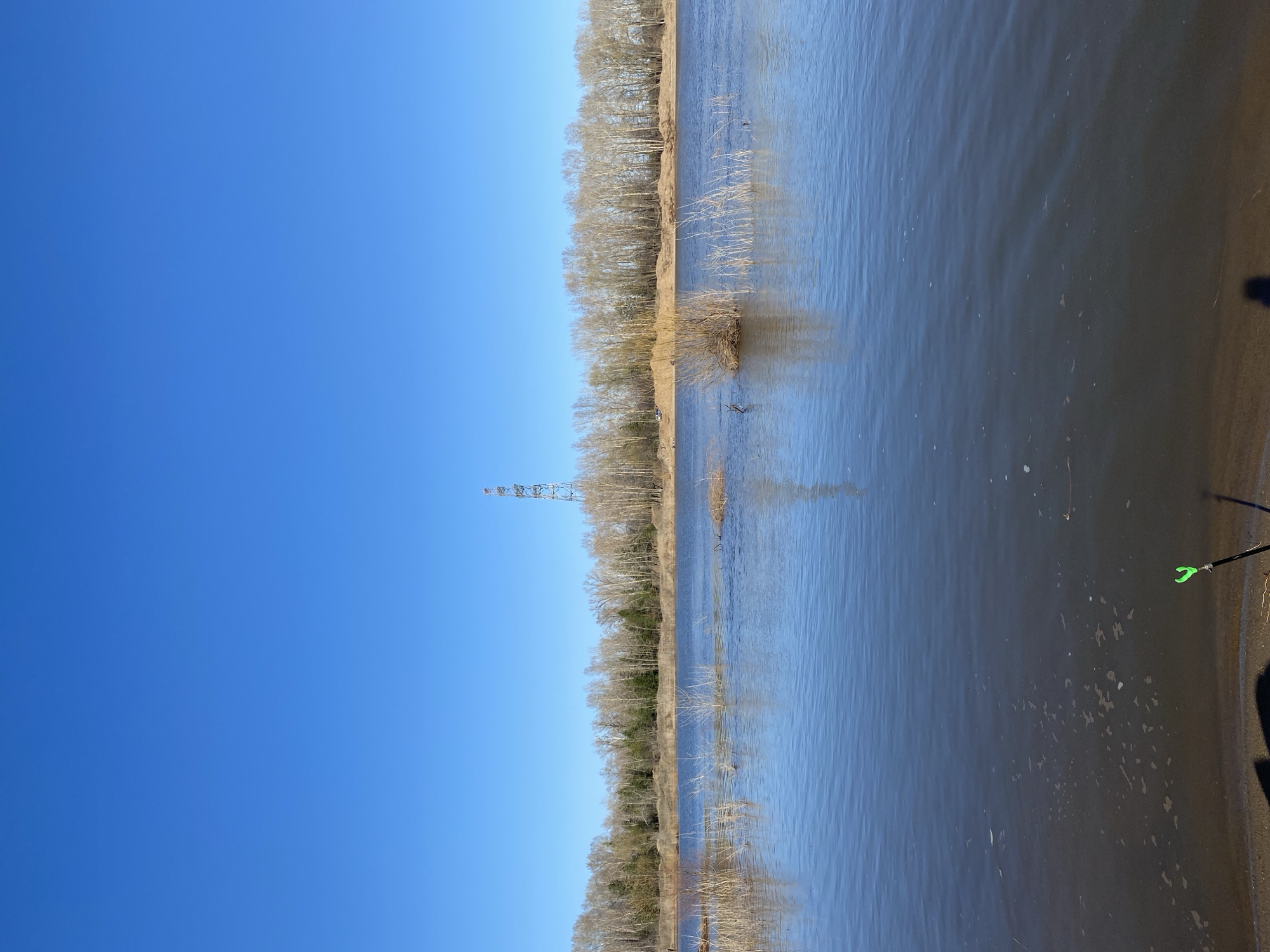The image depicts a landscape featuring a body of water, likely a river or lake, with a treeline along the opposite bank. In the distance, an electrical transmission tower is visible. The sky is clear and blue. The water's surface shows ripples and reflections. On the right edge of the frame, there is a portion of land and fishing equipment, including a rod holder. The location is in the city of Noviy, Russia.