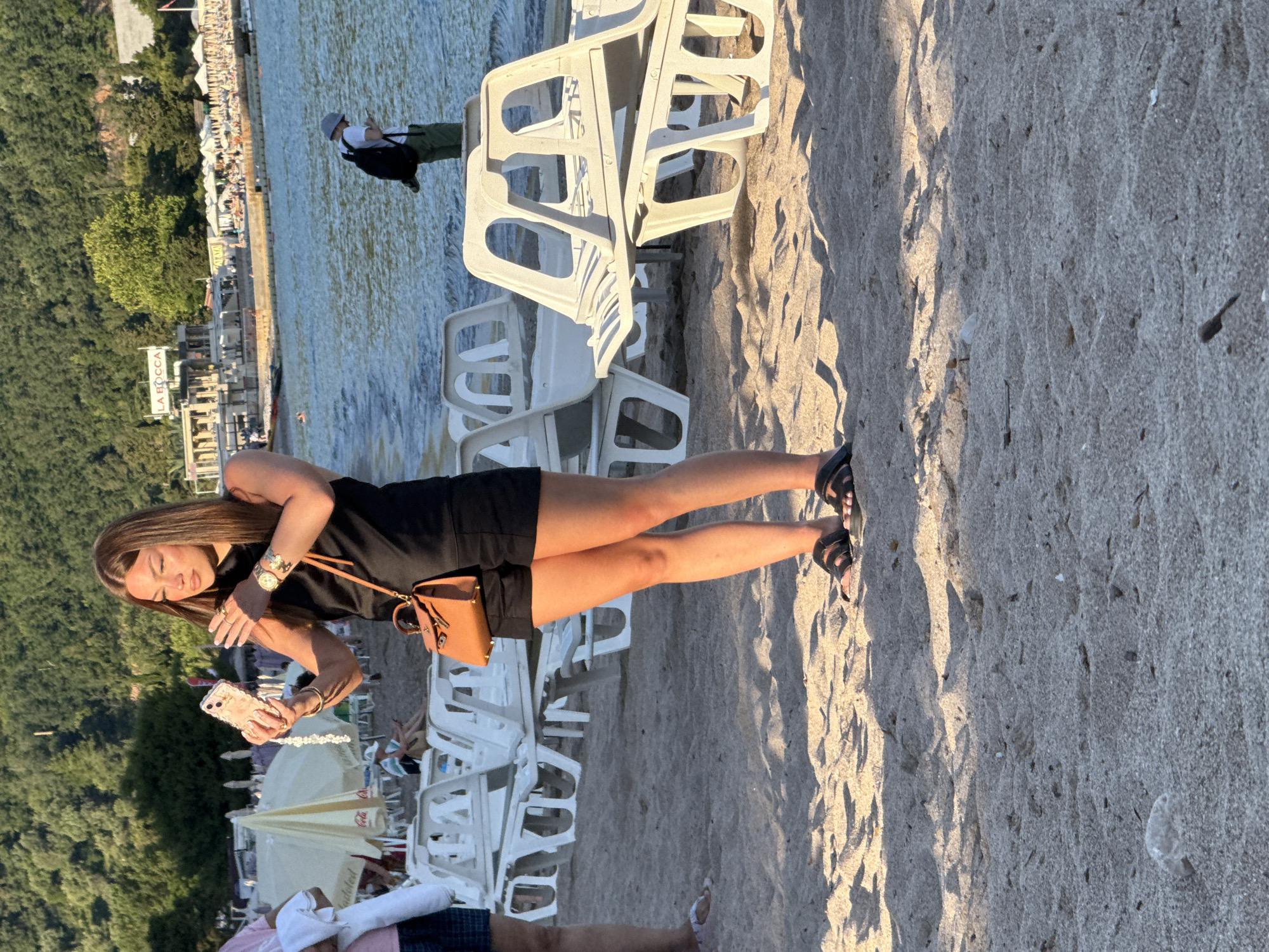 A young woman with long brown hair, wearing a black sleeveless top and black shorts, is standing on a sandy beach in Budva, Montenegro. She is holding a smartphone in her left hand, seemingly taking a selfie, and has a small brown handbag with a gold clasp hanging from her right shoulder. She is wearing a gold watch on her left wrist and dark sandals on her feet. Behind her, the beach is lined with white lounge chairs, some stacked. The sea is visible in the background, with a few people swimming. Further back, there is a waterfront promenade with buildings and umbrellas. The overall scene suggests a sunny day at a beach resort.
