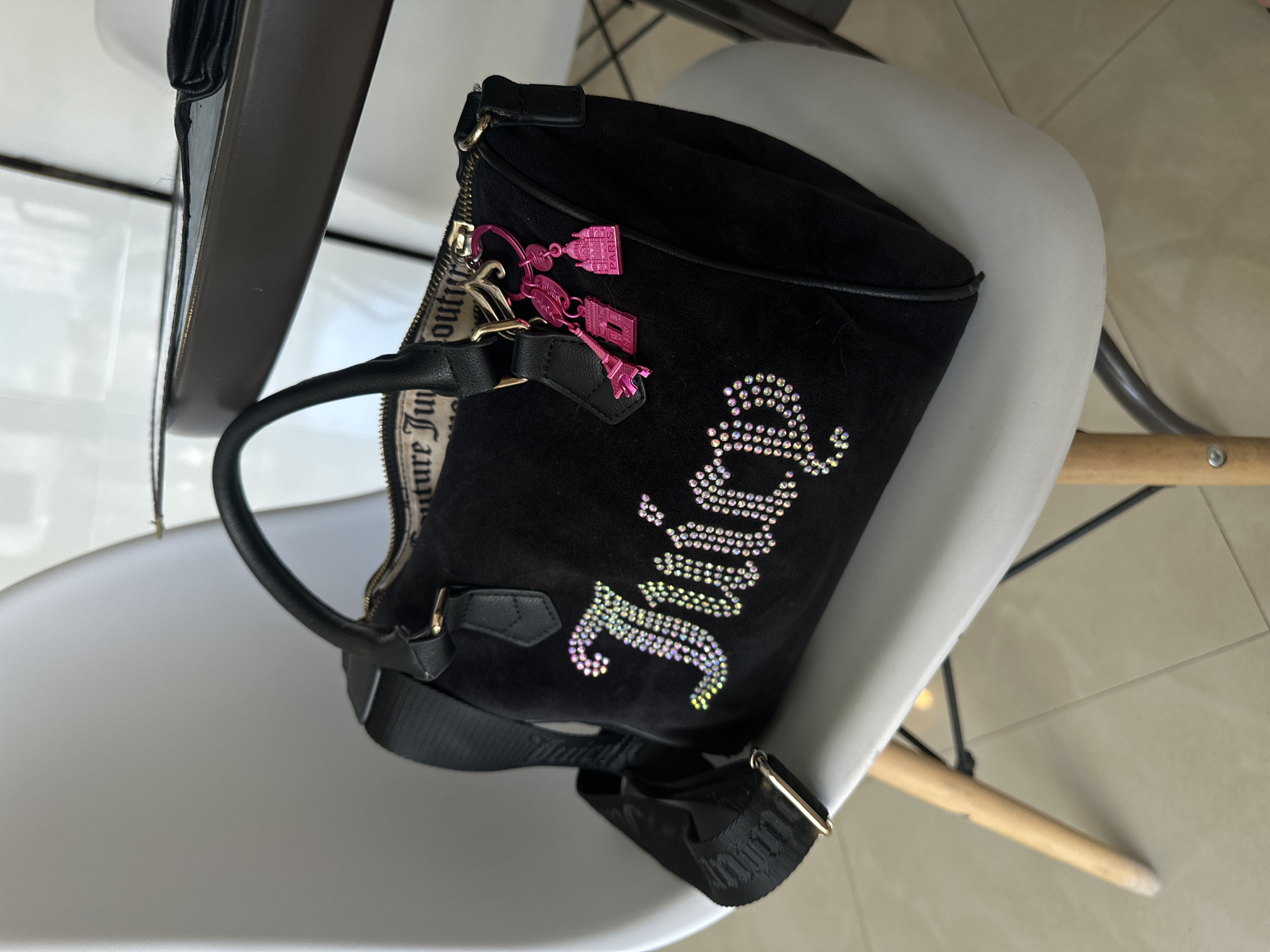 A black velour handbag with "Juicy" spelled out in rhinestones is placed on a white chair. The bag has a zipper closure and a strap. Two pink Eiffel Tower-shaped charms are attached to the zipper pull. The chair has wooden legs and is positioned in front of a dark-colored table. The background appears to be an indoor setting with light-colored flooring. There are no people visible in the image. There is no information in the image that suggests a location in Budva, Montenegro.