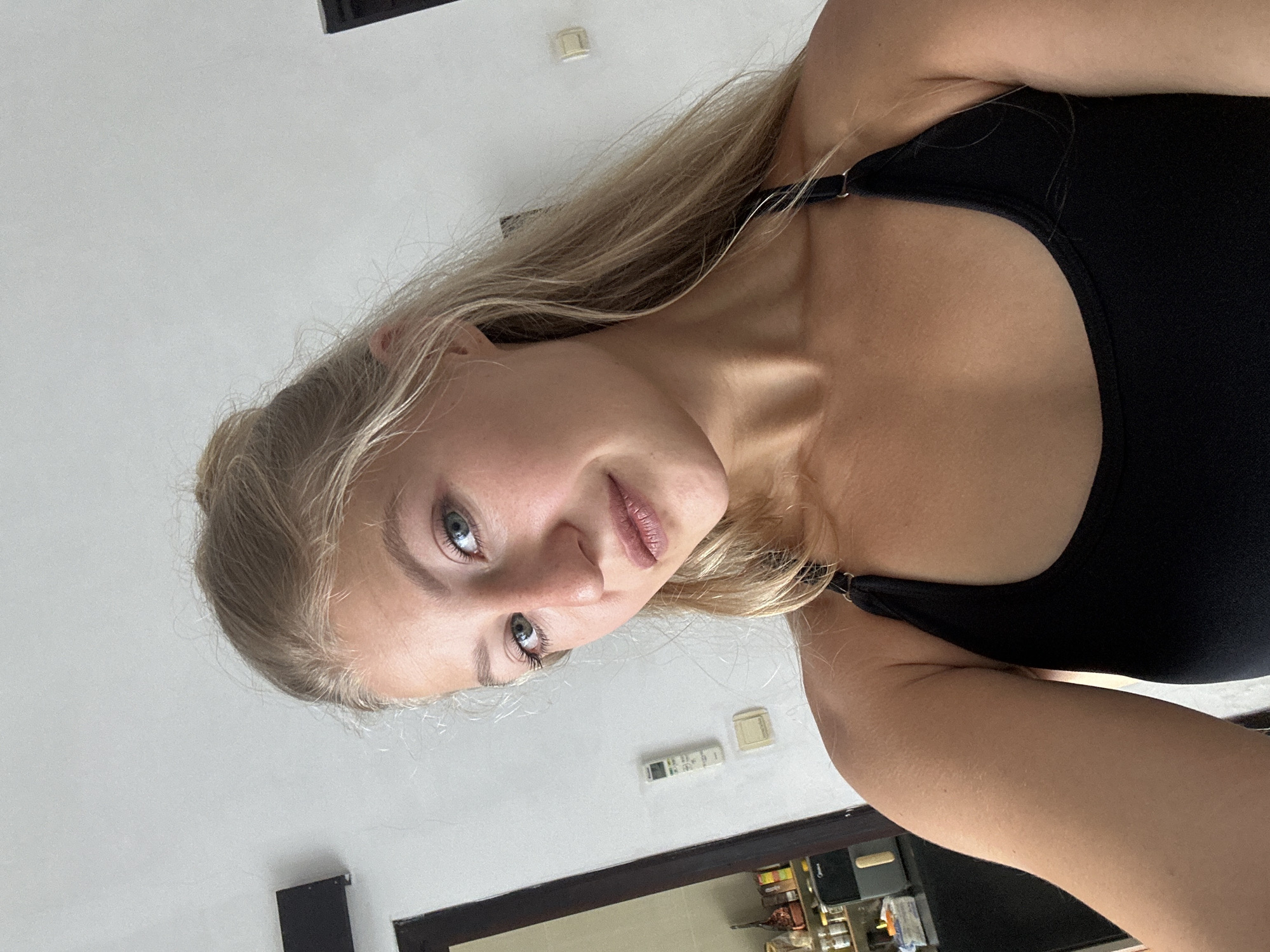 The image, rotated 90 degrees clockwise, captures a close-up portrait of a young woman in an indoor setting. She has fair skin, long blonde hair, and striking light blue eyes, looking directly towards the viewer with a neutral expression and slightly parted lips. She is wearing a black tank top or sports bra.
The background primarily features a plain white wall, illuminated by what appears to be natural daylight. A white air conditioning remote control and another small, rectangular white fixture are visible on the wall. Below these, a dark-framed mirror or window reflects part of an adjacent room, revealing shelves holding various jars and containers, suggesting a kitchen or storage space. The woman's right arm is partially visible along the lower edge of the frame. The scene is set in Kuta Selatan, Indonesia. No readable text is discernible on any visible objects.