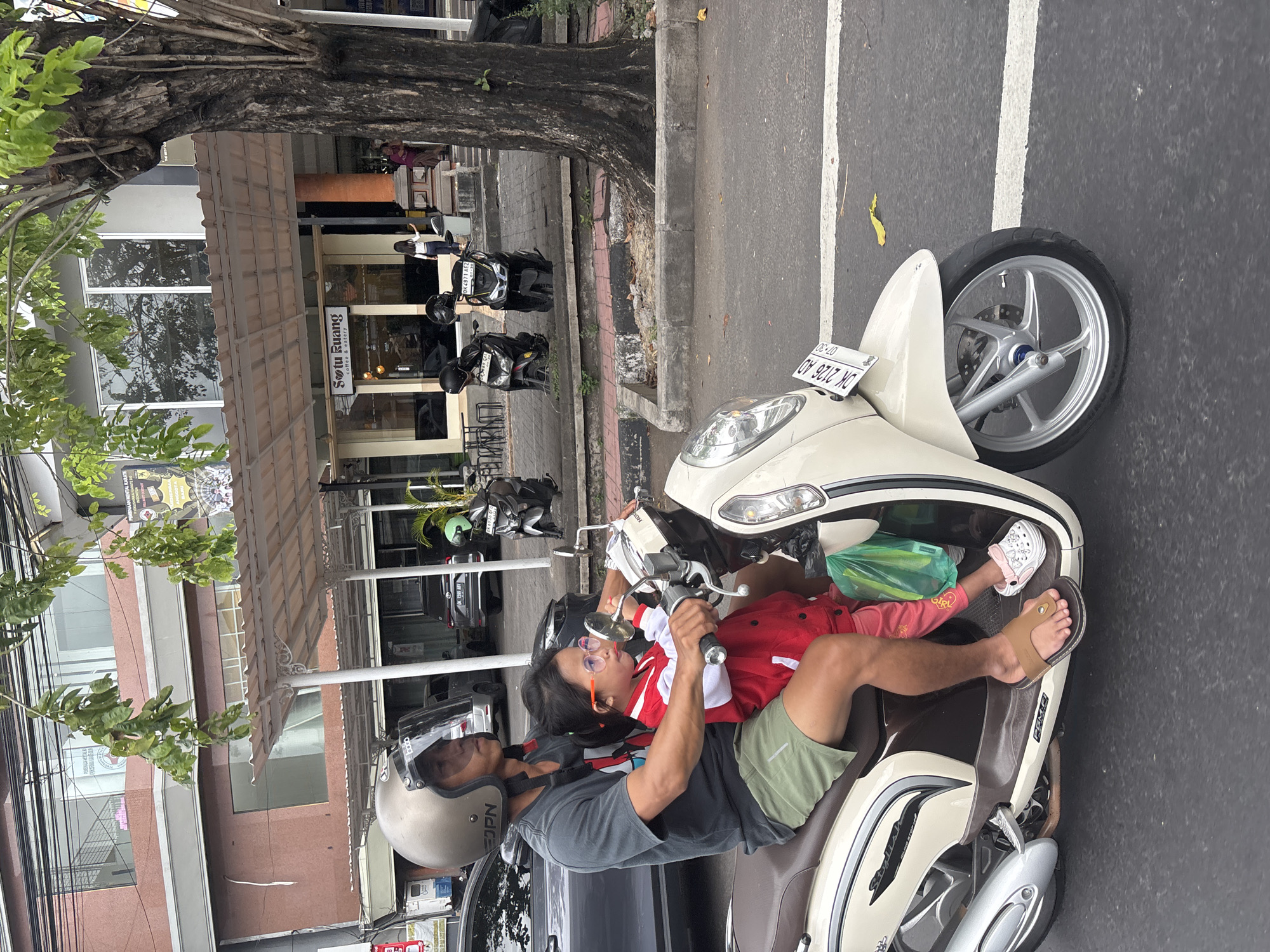 A man and a young girl are riding a cream-colored scooter on a street in Denpasar, Indonesia. The man is wearing a gray t-shirt, green shorts, a silver helmet, and brown sandals. The girl is wearing a red and white jacket, pink pants, and white clogs. She is holding onto the handlebars and appears to be looking to the right. A green plastic bag is hanging from the back of the scooter. In the background, there are buildings, trees, and other scooters parked along the sidewalk. The street is paved and there is a white line marking a lane. The overall scene depicts a typical day on a busy street in a city.