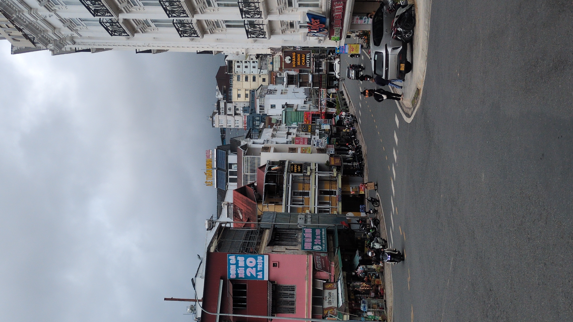 The image depicts a street scene in Da Lat, Vietnam, characterized by its hilly terrain and architecture typical of the city. On the left, a pink building with red trim features signage advertising "COM GA XOT MO" and "LIEN THANH." Adjacent to it are multi-story buildings with balconies and various commercial establishments, including shops and restaurants, indicated by signs like "PHUC LOC." The street is paved and marked with white dashed lines. A number of motorcycles are parked along the sidewalk and on the road.
In the center of the image, a silver SUV is parked on the right side of the road. Two individuals are standing near the SUV, one wearing a black jacket and the other in checkered pants. Further down the street, several motorcycles are in motion, and more are parked. Buildings of varying heights line both sides of the street, with a prominent white building on the far right with ornate balconies. Signs for "HOANG MINH CHAU HOTEL" are visible. The sky is overcast with grey clouds. The overall impression is of a busy urban environment, likely a commercial district in Da Lat.