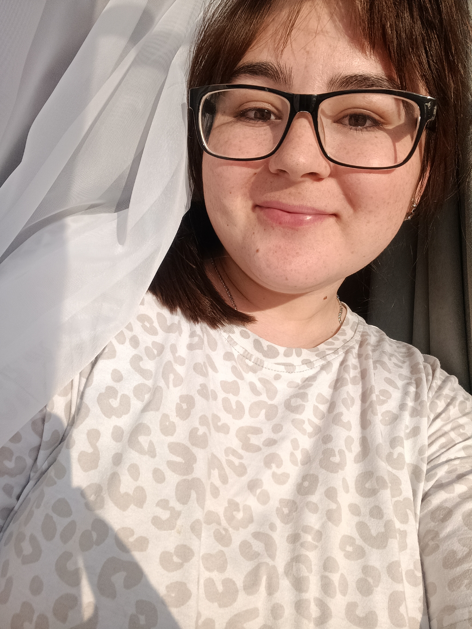 An adult female is featured in a close-up portrait, located indoors in the city of Noviy, Russia. The subject has medium-brown hair with bangs, wears black-framed eyeglasses, and displays light skin with visible freckles. Her brown eyes are open and directed forward, and she exhibits a faint, closed-mouth smile. She is wearing an off-white t-shirt with a light beige leopard print pattern. A thin silver-colored chain necklace is visible around her neck, and a small silver-colored hoop earring is present in her right ear. The scene is illuminated by natural light originating from the left, creating highlights on the subject's face and glasses, and casting subtle shadows on her clothing. A sheer, white, pleated curtain is draped on the left side of the frame, partially obscuring the subject's left side. A darker, solid-colored curtain, likely gray, is visible in the background on the right side. The subject is actively posing for the photograph.