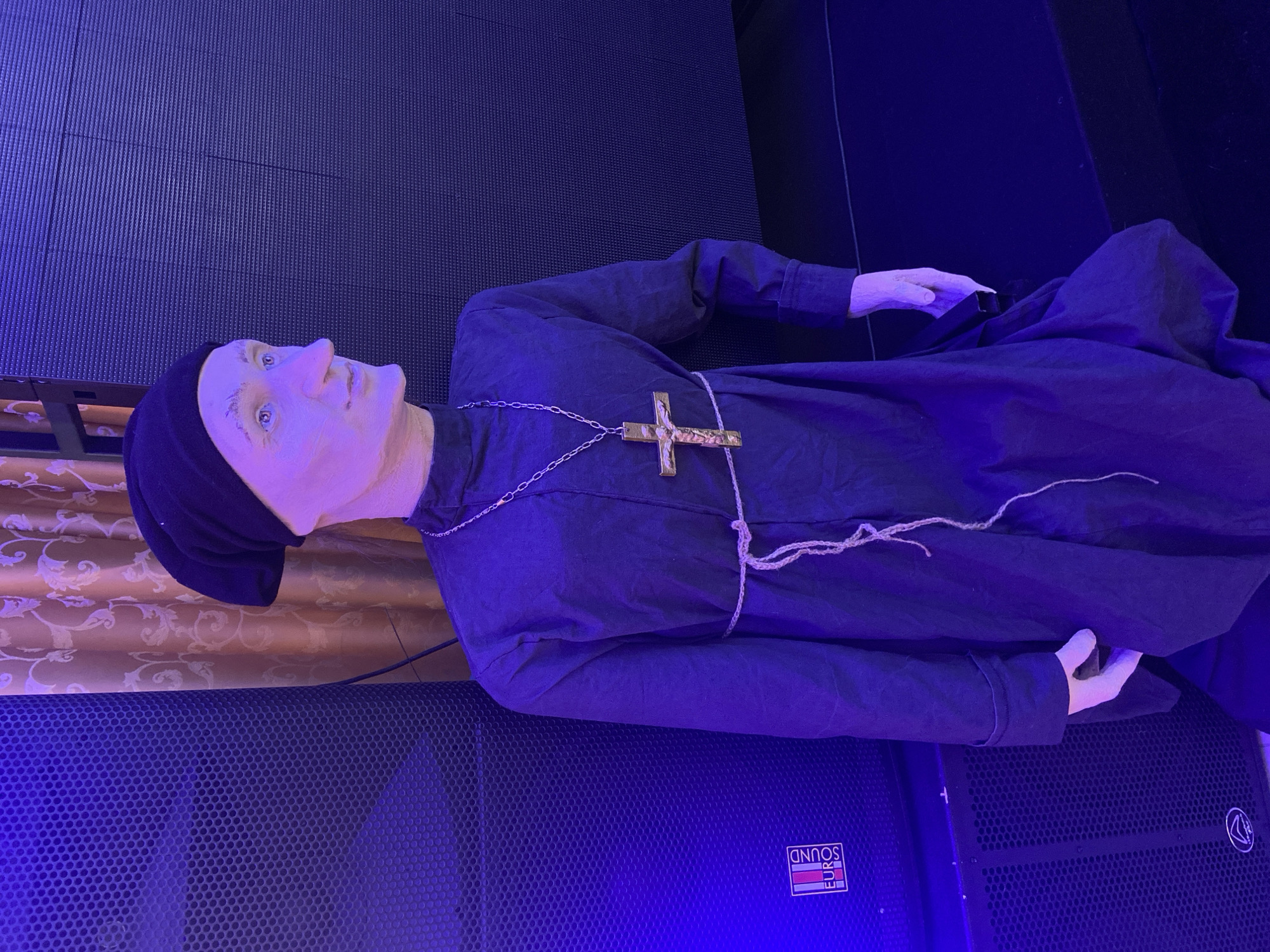 The image shows a mannequin dressed as a monk lying on top of a large speaker cabinet with the brand "EURO SOUND" visible. The mannequin wears a black robe, a black cap, and a large, golden cross on a chain. The mannequin's skin is pale and its features are exaggerated. The speaker cabinet is black and has a perforated front. The background is a mix of surfaces, including a patterned fabric and what appears to be a black, possibly corrugated surface. The scene is lit with a blue light. The city of Chaikovsky, Russia, is not discernible from the image.