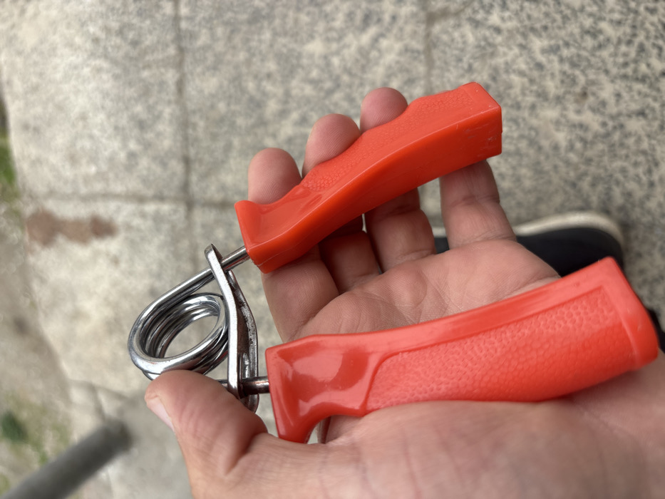The image displays a human hand, exhibiting light skin pigmentation, holding a red and silver hand gripper. The hand gripper consists of two red, ergonomic, injection-molded plastic handles, one featuring a textured grip area, connected by a coiled chrome-plated steel spring and two steel pins. The device is held in a relaxed, open position. The background is out of focus, revealing a light grey paved surface, consistent with concrete or flagstones, marked by visible seams. A blurred, vertical metallic pole is discernible in the upper-left background, and a dark, indistinct object, possibly part of a shoe, is partially visible beneath the hand on the right. The scene is situated outdoors in the city of Lehvaz, Armenia.