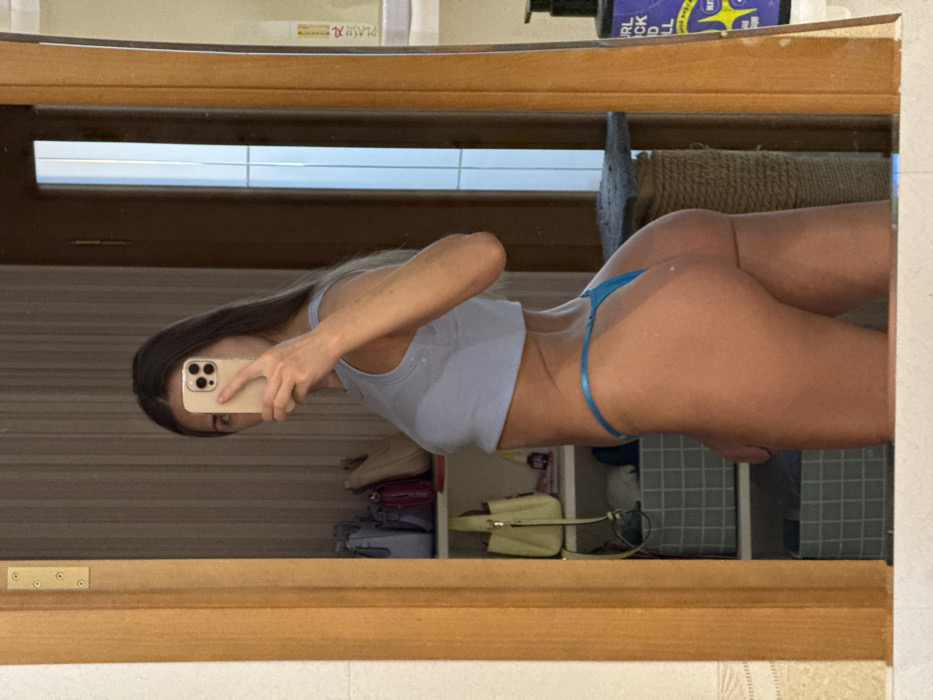 A woman in a light blue crop top and a blue thong stands facing left, holding her cell phone in front of her. She is reflected in a mirror. Behind her are shelves with various items, including shoes, purses, and containers. Part of a wooden frame is visible surrounding the mirror's reflection.