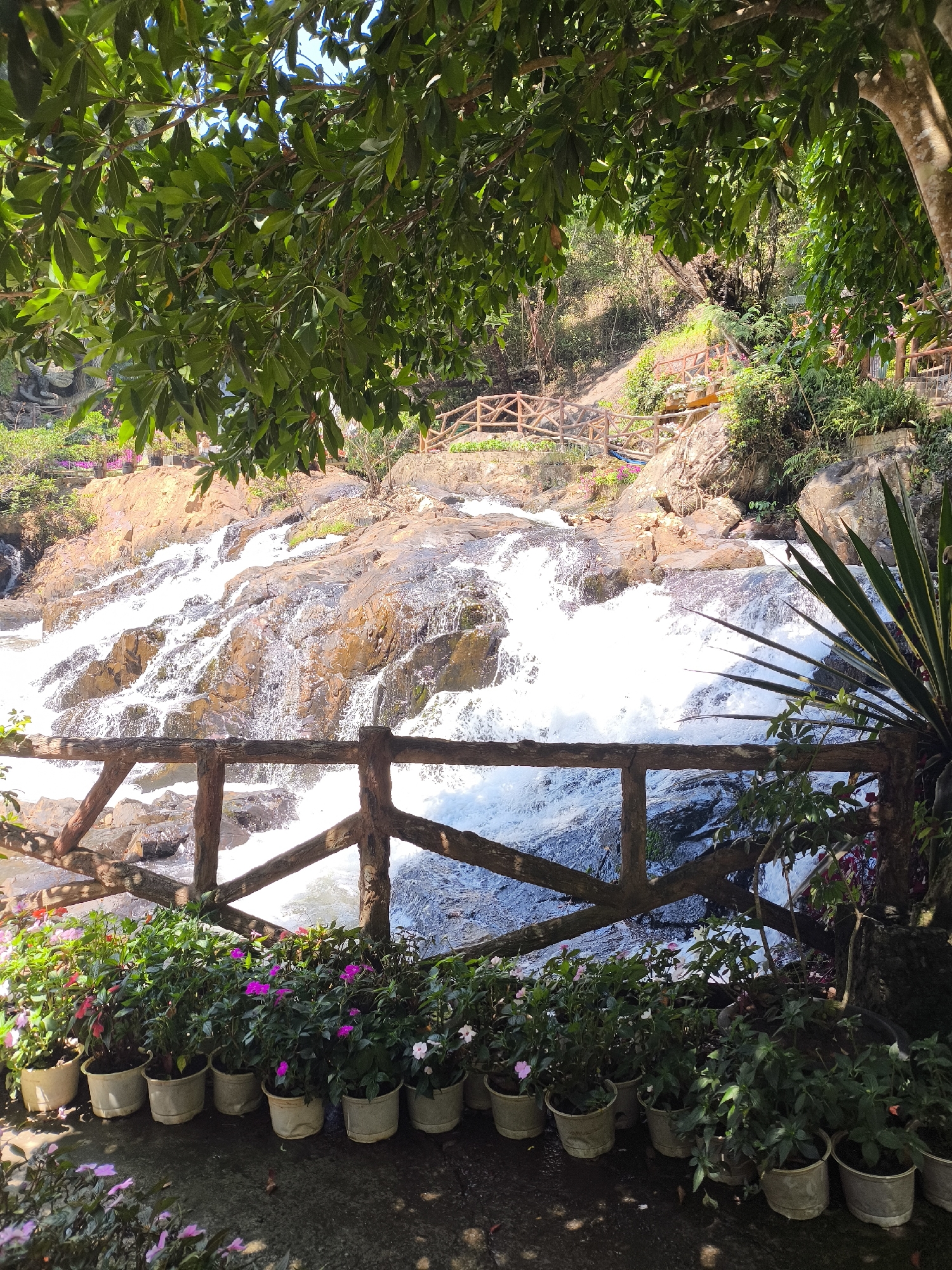 The image depicts a multi-tiered waterfall in a landscaped environment. The water cascades over a series of rocky ledges. A rustic wooden fence runs along the foreground, lined with numerous potted plants bearing colorful flowers. Lush green foliage, including trees and various plants, frames the top and sides of the scene. The location is in Da Lat, Vietnam, as evidenced by the landscape. The scene is observed in daylight.