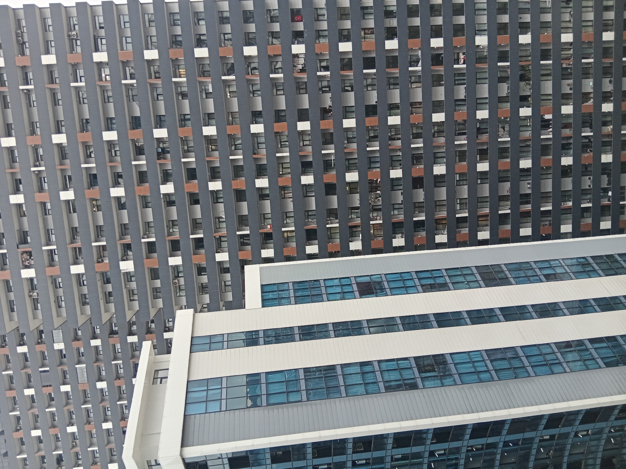 The image shows a high-angle view of two buildings in Batumi, Georgia. The building in the background is a tall residential structure with a repeating pattern of windows and gray vertical columns. Some windows have glimpses of interiors, including a red flag in one. The building in the foreground is a lower office building with white horizontal sections and blue-tinted windows. The overall environment seems urban and dense. The scene is viewed from above. There are no clearly discernible people. The focus is on the architectural elements of the buildings.