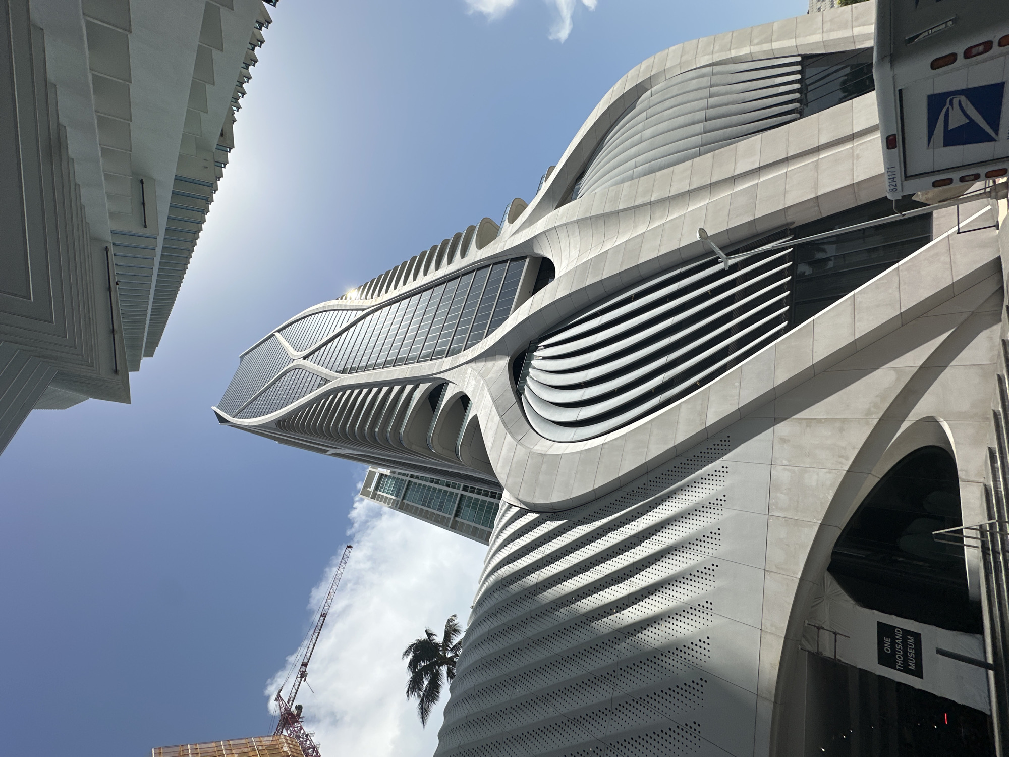 A low-angle, wide shot captures the exterior of the One Thousand Museum building in Miami, Florida, on a clear, sunny day. The avant-garde skyscraper dominates the frame with its sculptural, fluid concrete facade and glass curtain walls. Shadows from the building's distinctive curves and the sun's rays create a dramatic interplay of light and dark across its surfaces.
In the foreground, a sign for "ONE THOUSAND MUSEUM" is visible near the entrance, set against a curved, perforated concrete wall. Stairs lead up to the building's entrance. To the left, a construction crane extends into the bright blue sky, indicating ongoing development in the area. A palm tree with windblown fronds adds a tropical element to the scene.
Further back, another modern skyscraper with a patterned facade can be seen. The sky is a vibrant blue with a few scattered white clouds. A white United States Postal Service van is parked in front of the building. The architecture of the One Thousand Museum is characterized by its organic, flowing lines, designed by the late architect Zaha Hadid.