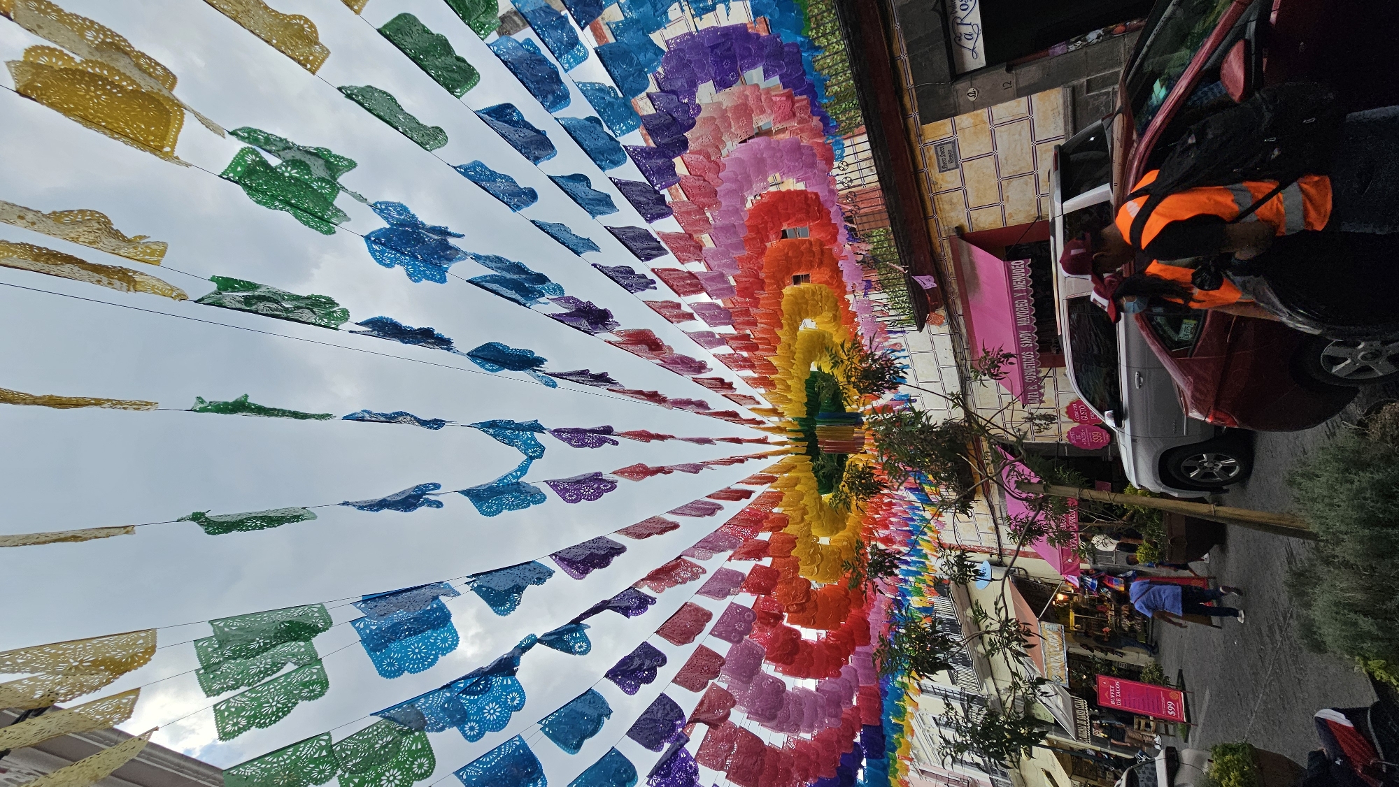 This vibrant street scene, likely in Puebla, Mexico, is dominated by an elaborate overhead display of "papel picado" banners. Hundreds of these traditional Mexican cut-paper decorations, strung on wires, create a colorful canopy in a rainbow gradient. Starting with yellows, greens, and blues on the left, the colors transition through purples, reds, oranges, and pinks towards the right, creating a tunnel-like effect along the thoroughfare. The intricate patterns of the individual banners are visible against a cloudy, overcast sky.
Below the festive decorations, the street is lined with buildings featuring various storefronts. On the right, a red car is parked, with a person wearing an orange high-visibility vest leaning into it, appearing to be attending to something or someone. Further down the street, a silver SUV is parked, and a few pedestrians are visible walking on the pavement. The buildings exhibit different architectural styles and colors, including one with pink awnings and a tiled facade. Lush green foliage from small trees and plants spills from planters along the sidewalk.
Visible text on the storefronts includes "$99 BUFFET DE TACOS" on a pink sign, and "AURA B. COSMETICOS SANDY MAYOREO Y MENUDEO" on another pink awning, suggesting a cosmetics or general store. Part of a sign reads "Come GUSTO" and another "La Ros...". A number "12" is also visible on a building facade. The general atmosphere is lively and celebratory, indicative of a special occasion or simply a culturally rich urban environment. The time of day appears to be daytime, possibly with indirect sunlight due to the cloud cover.