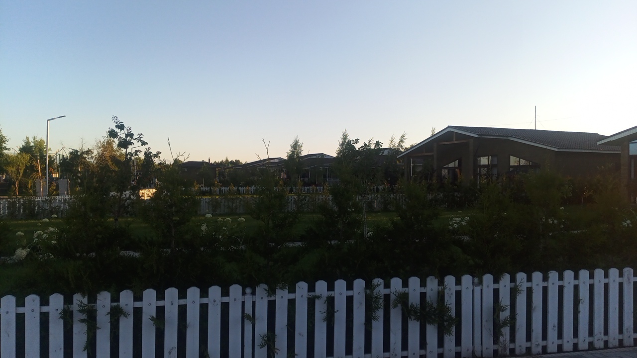 The image depicts a residential area in Choktal, Kyrgyzstan, under a clear sky. In the foreground, a white picket fence runs horizontally across the frame, partially obscuring a lush green lawn dotted with white flowers. Behind the fence, various trees and shrubs create a dense layer of foliage. In the middle ground, a row of modern houses with dark roofs are visible, separated by more white picket fences and trees. A streetlamp stands to the left of the houses, casting a faint light. The overall impression is one of a quiet, suburban landscape during the daytime.