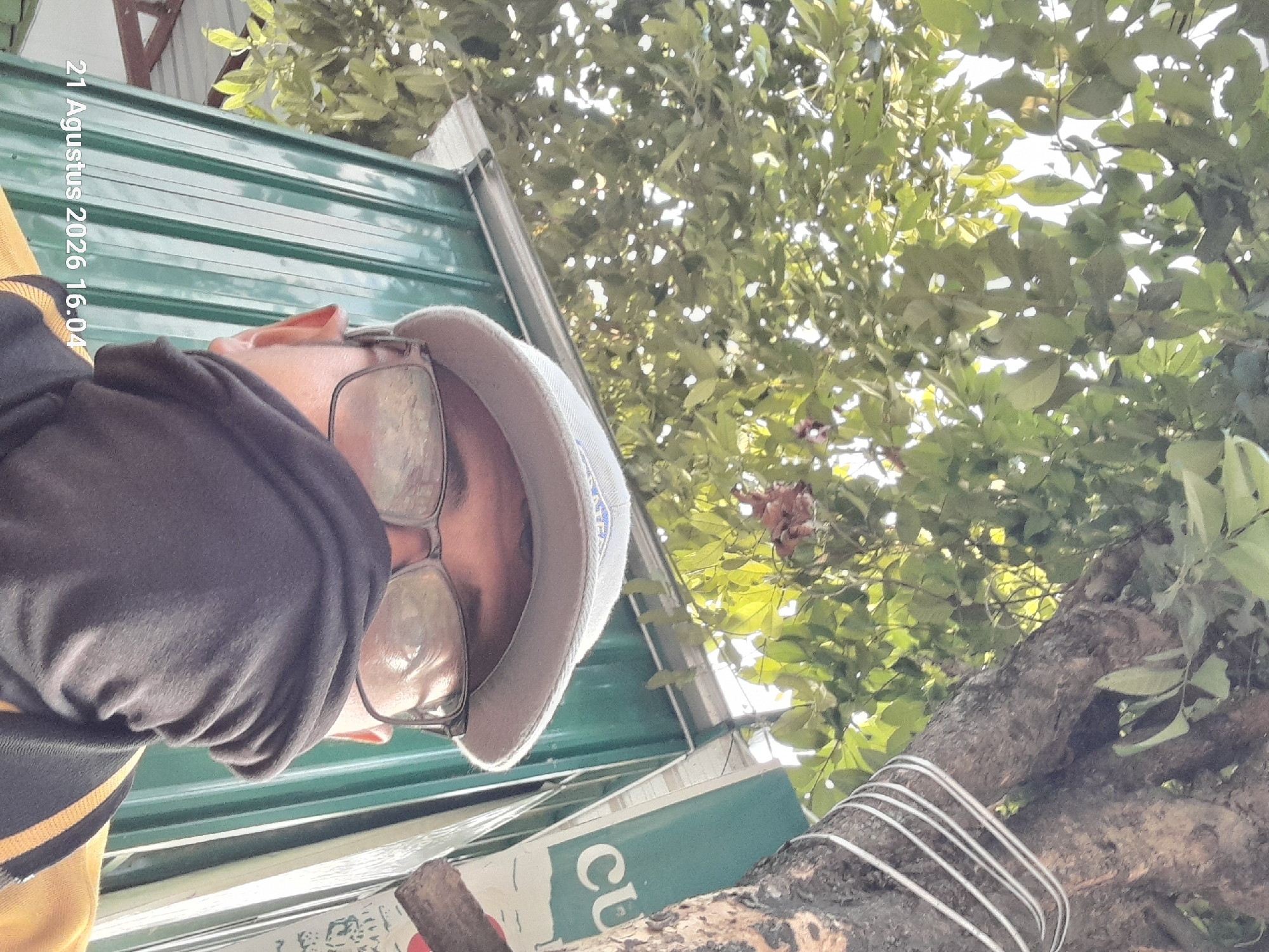 The image is a selfie taken outdoors, from a low angle looking up at the subject and the environment. The subject is wearing glasses, a light-colored baseball cap, and a black face covering that obscures their mouth and nose. They are also wearing a yellow shirt with black trim. The background consists of a large tree with green leaves and a corrugated metal wall that appears to be part of a building. The wall is painted a muted green color, and there is a white sign with the letters "CU" visible on it. The image is dated "21 Agustus 2026 16.04". The date suggests the location is in Indonesia, and Takeran is a town in the East Java province of Indonesia.