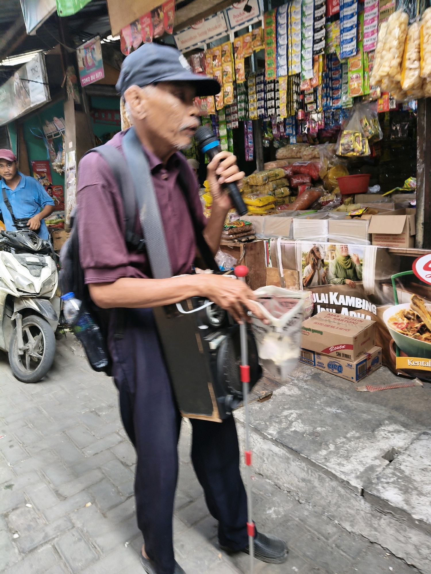 A man in a dark blue baseball cap and a maroon polo shirt is walking on a cobblestone street in front of a shop in Setiamekar, Indonesia. He is holding a microphone in his right hand and a cane with red accents in his left. He has a backpack on his shoulders. A white motorcycle is parked to his left. The shop behind him is stocked with various food items, snacks, and household goods. A poster with images of people and text in Indonesian is visible on the shop's facade.