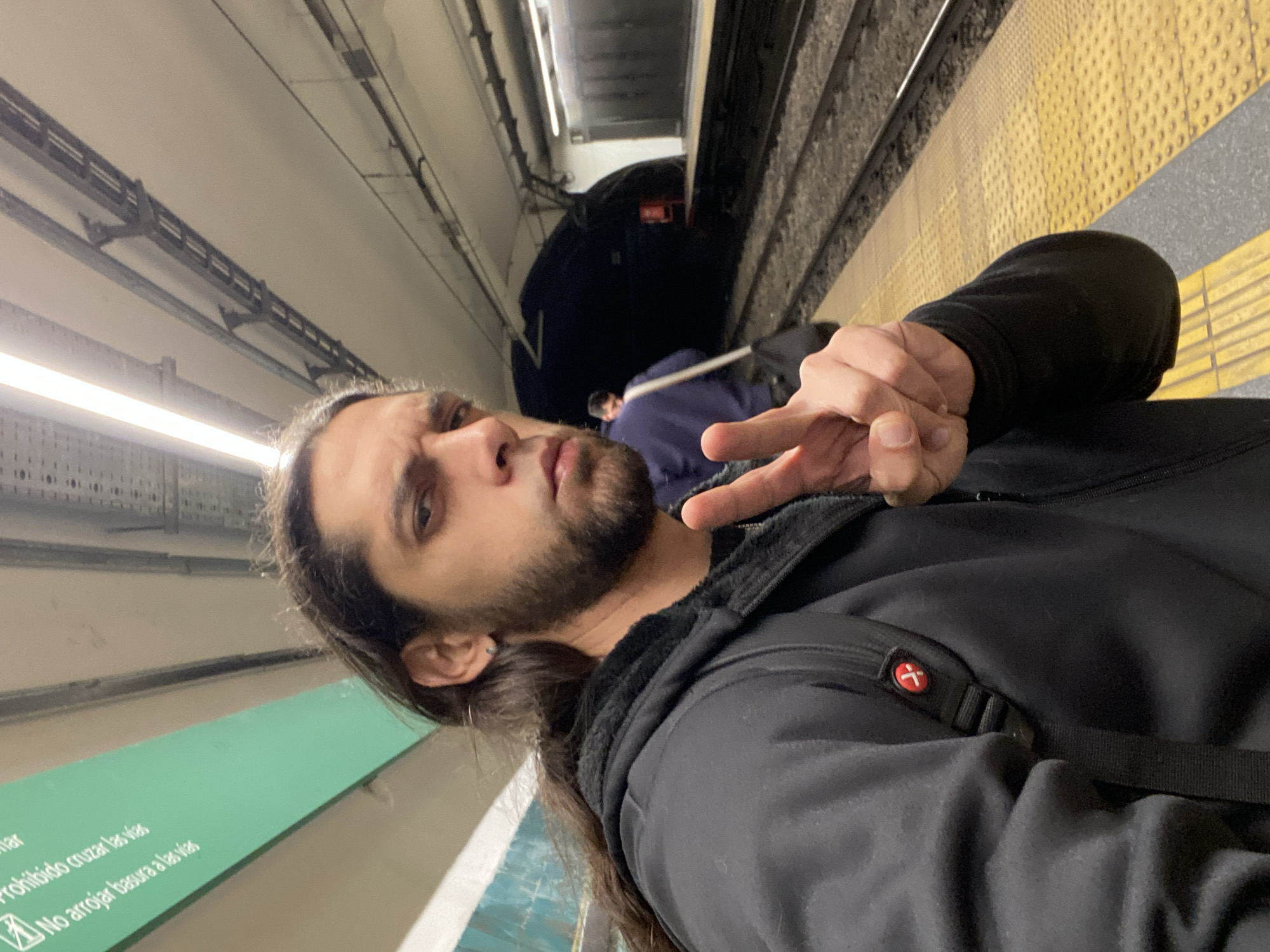 The image shows a man with long hair and a beard, wearing a black jacket, standing on a subway platform. He is making a peace sign with his hand. Behind him is a subway train in the tunnel and another person in a blue jacket. The platform has yellow tactile paving. A sign in Spanish is partially visible on the wall. The location is the Buenos Aires, Argentina subway.