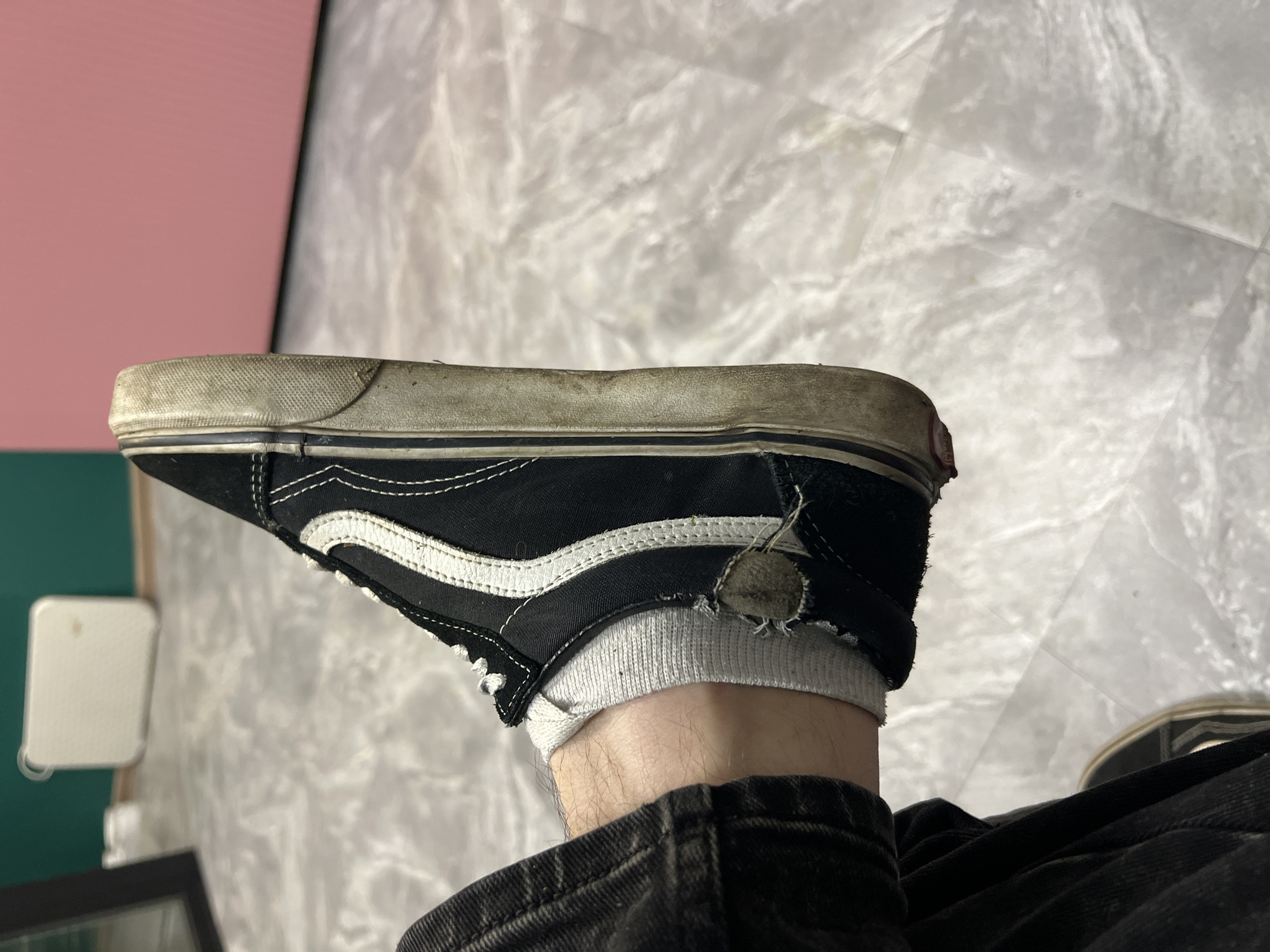 The image shows a close-up, side-view of a black and white Vans sneaker on a person's foot. The sneaker is worn, with scuff marks and dirt on the sole and sides. The white stripe on the side is slightly discolored. The white sock is visible above the shoe and has a tear near the ankle. The person is wearing dark, possibly denim, pants. The background appears to be an indoor setting with a floor that has a marble-like pattern. In the background, there are blurry objects that suggest furniture or appliances, and a wall painted in dark green and a lighter pinkish-red. The image does not provide any information about the location in the city of Ленинский, Russia.