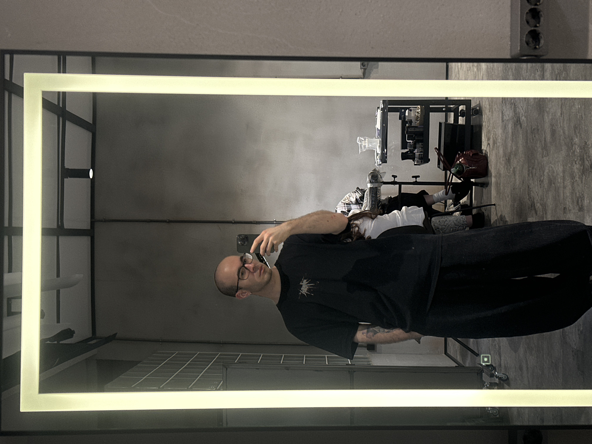 A person is reflected in a large, rectangular mirror with a bright light frame. The person, likely male, is bald, wearing glasses, a dark t-shirt with a design, and dark pants. He is holding a phone in his hand as if taking a picture of himself in the mirror.
The background suggests a studio or workshop setting. There is metal framework visible on the left side, a metal cart with various equipment on the right, and a gray wall behind the cart. A second person is partially visible behind the subject and is sitting. The environment appears to be indoors with a modern industrial aesthetic. The image does not contain enough information to determine if the scene is in Moscow, Russia.