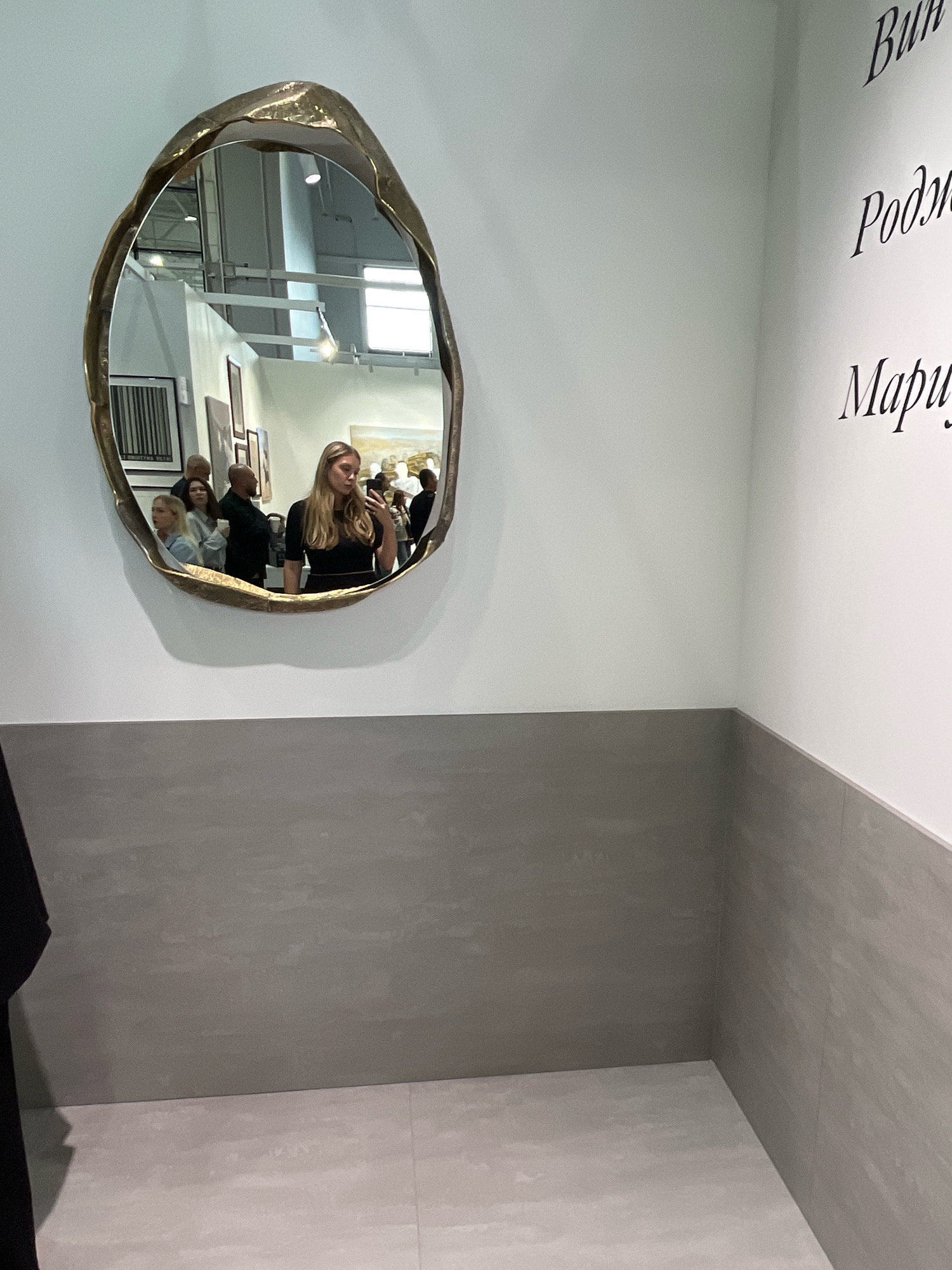 In an interior space, a woman with blonde hair is taking a selfie in a uniquely shaped mirror with a gold frame. She is wearing a black top. Reflected in the mirror are other people, some looking at art on the walls, and a painting. The walls are painted white and there is a dark gray tiled wainscoting. In the foreground, a black garment is hanging from the left side of the frame. On the wall to the right, there are handwritten words in black calligraphy: "Вик" (Vik), "Рожд" (Rozhd), and "Мариу" (Mariu). This scene is located at an art exhibition or gallery, likely within the city of Khimki, Russia, given the Cyrillic script and typical exhibition aesthetics.
