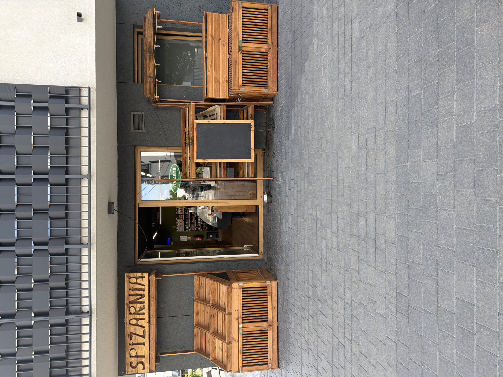 The image shows the exterior of a shop called "SPiZARNiA" in an unknown city. The shop has a dark grey textured facade. Above the entrance, there is a wooden sign with the shop's name carved into it. To the left of the entrance, there is a wooden display stand with multiple compartments. To the right of the entrance, there is another wooden structure with a canopy and a chest-like base. Inside the shop, visible through the glass doors, are shelves stocked with products and a refrigerated display case. In front of the shop, on the paved ground, is a wooden easel with a blackboard. A person is partially visible in the reflection of the shop window, holding a phone. The ground is paved with grey rectangular tiles.