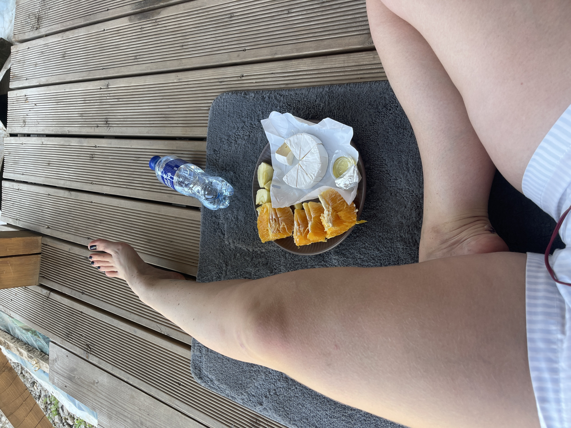 A person is lying on a gray towel on a wooden deck. A plate of cheese, orange slices, and a small container of an unidentified food item is on the towel near the person's legs. A water bottle is on the deck. The person's left arm extends towards the left side of the frame, with their foot visible on the deck. A building or other structure is visible on the left side. The location is likely outdoors, suggested by the wooden deck and the partial view of the surroundings. The location in the city of Мосметаллоконструкция Онт, Russia, is undetermined.