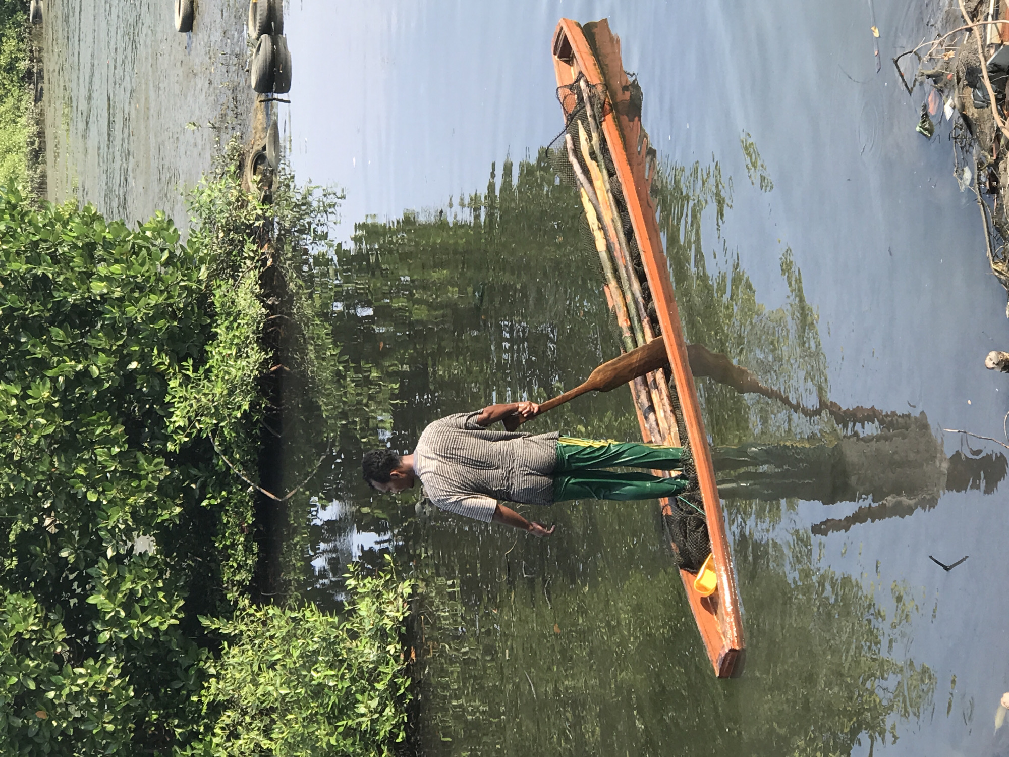 A man stands on a narrow, rustic, wooden boat in a body of water. The boat is laden with sticks and a net. He is using a long oar to propel the boat. The man has dark hair and is wearing a horizontally striped, collared shirt and bright green pants. Lush green foliage lines the left bank of the waterway. The water surface reflects the surrounding trees and sky. Debris and tires are visible along the right bank, indicating pollution. The location is Lhokseumawe, Indonesia.