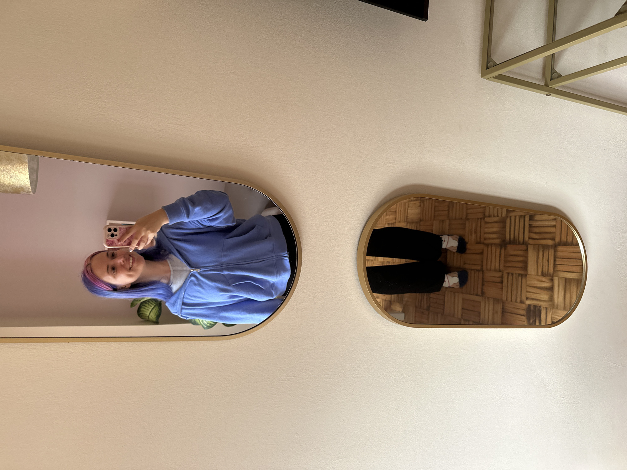 A woman takes a selfie in a mirror. She wears a blue zip-up hoodie and has dyed purple and blue hair. She stands in front of a white wall, where two rounded mirrors hang. One mirror reflects the woman, and the other reflects a hardwood floor and black pants. A portion of a plant is visible in the woman's mirror. A gold shelf is at the top right of the frame. The location is likely an indoor setting, possibly in Gijón/Xixón, Spain.