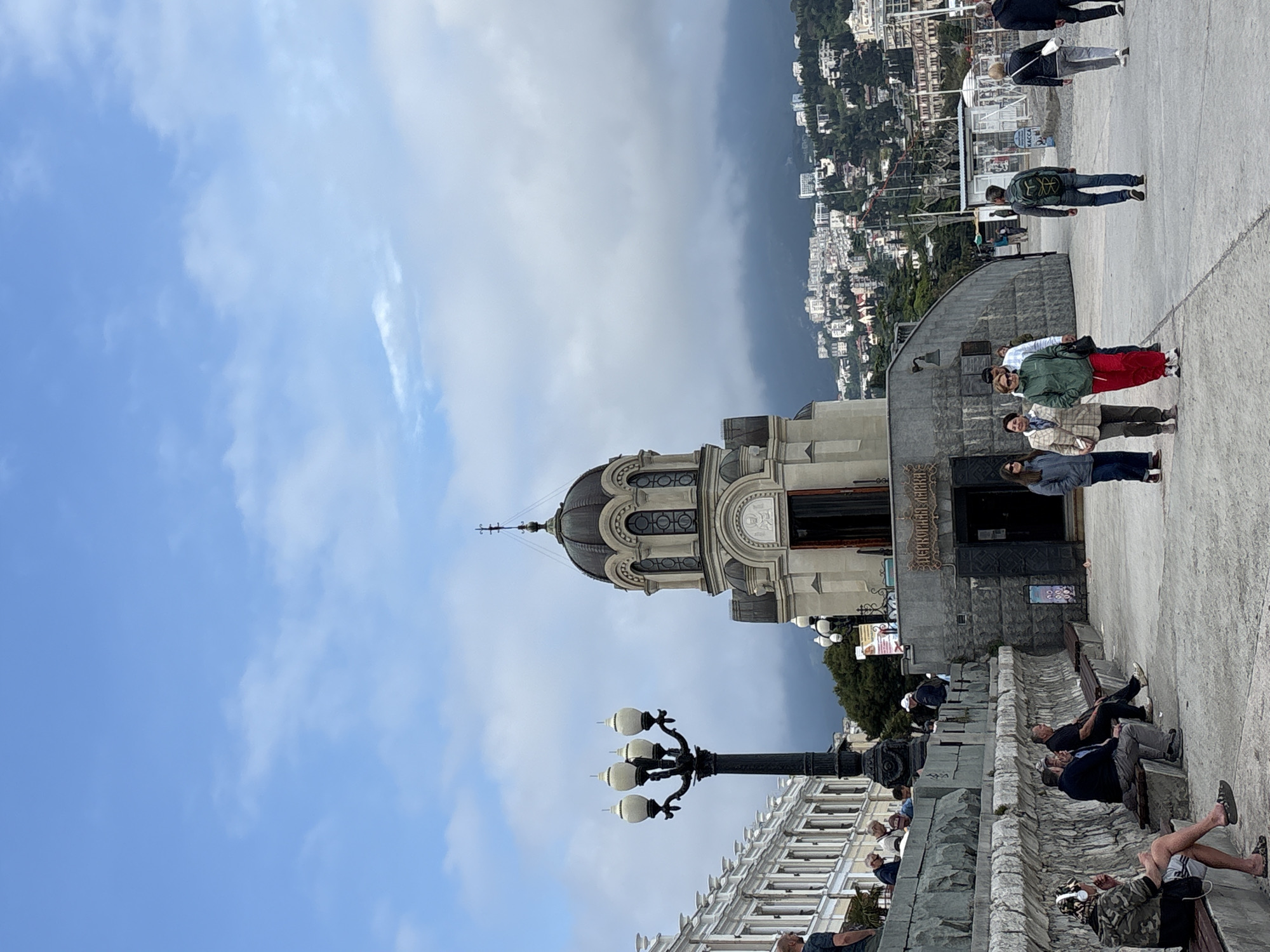 The image depicts a scene in the city of Yalta, Ukraine. A church or cathedral with a prominent dome is a central feature, constructed from light-colored stone. A dark, ornate lamppost stands in front of the building. A group of people are standing near the entrance of the building. Further away, people are seen sitting on benches. In the background, a hillside dotted with buildings is visible under a sky with a mix of blue and cloud cover. The ground appears to be paved.