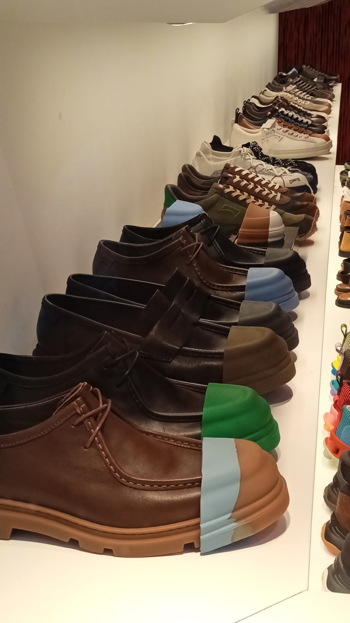 A display of men's shoes is shown on a white shelf. The shoes are arranged in rows, with the closest row featuring brown leather derby shoes. These shoes have visible stitching, laces, and thick tan soles. Further back, the display continues with several rows of various styles of men's shoes, including sneakers and casual shoes, in colors like brown, white, blue, and green. The shoes are placed on a white surface that recedes into the background. To the right, a vertical display shows a collection of colorful, sporty sneakers. The background is a plain white wall with a horizontal light fixture visible at the top. The overall scene depicts a retail environment, likely a shoe store, with the shoes presented in an organized and visually appealing manner. The image was taken in Milan, Italy.