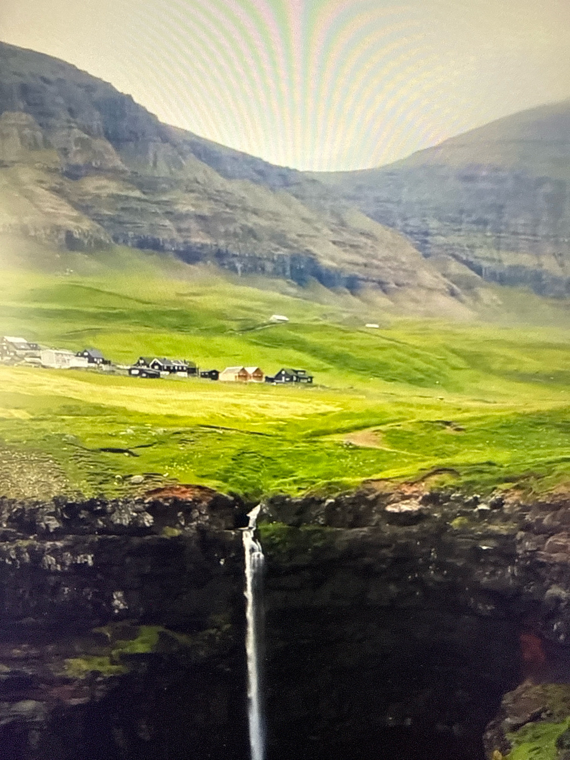 The image shows a dramatic landscape featuring a waterfall cascading from a cliff edge into a dark abyss below. The cliff is covered in dark rock and some green moss. Above the cliff, rolling green hills stretch towards a backdrop of majestic, layered mountains. A small cluster of houses with dark roofs and some colorful buildings are visible nestled in the green landscape in the middle ground. The sky is bright and appears to be illuminated by a soft, diffused light, possibly from the sun behind clouds or a haze. The scene evokes a sense of natural beauty and ruggedness. This image does not depict the city of Netanya, Israel.