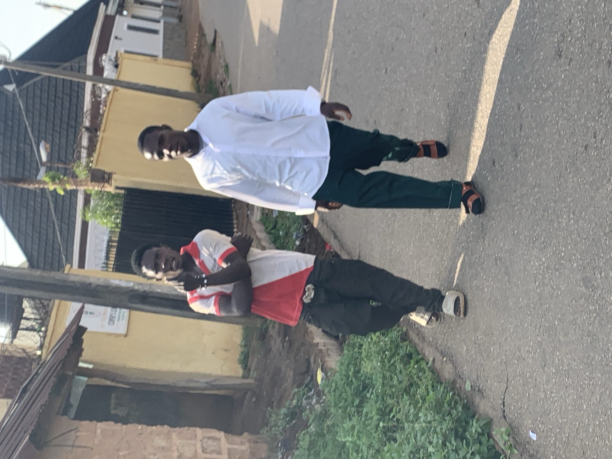 In Jos, Nigeria, on a paved street, two young men are walking. The man on the left, with dark hair and skin, wears a red and white polo shirt, black pants, and white sandals. He has his arms crossed, and he is looking to his left. The man on the right, also with dark hair and skin, is wearing a white long-sleeved shirt, green pants, and brown and black sandals. He is looking straight ahead as he walks. The environment appears to be a residential area, with buildings and vegetation visible in the background. The sun casts shadows on the street, indicating it is daytime.