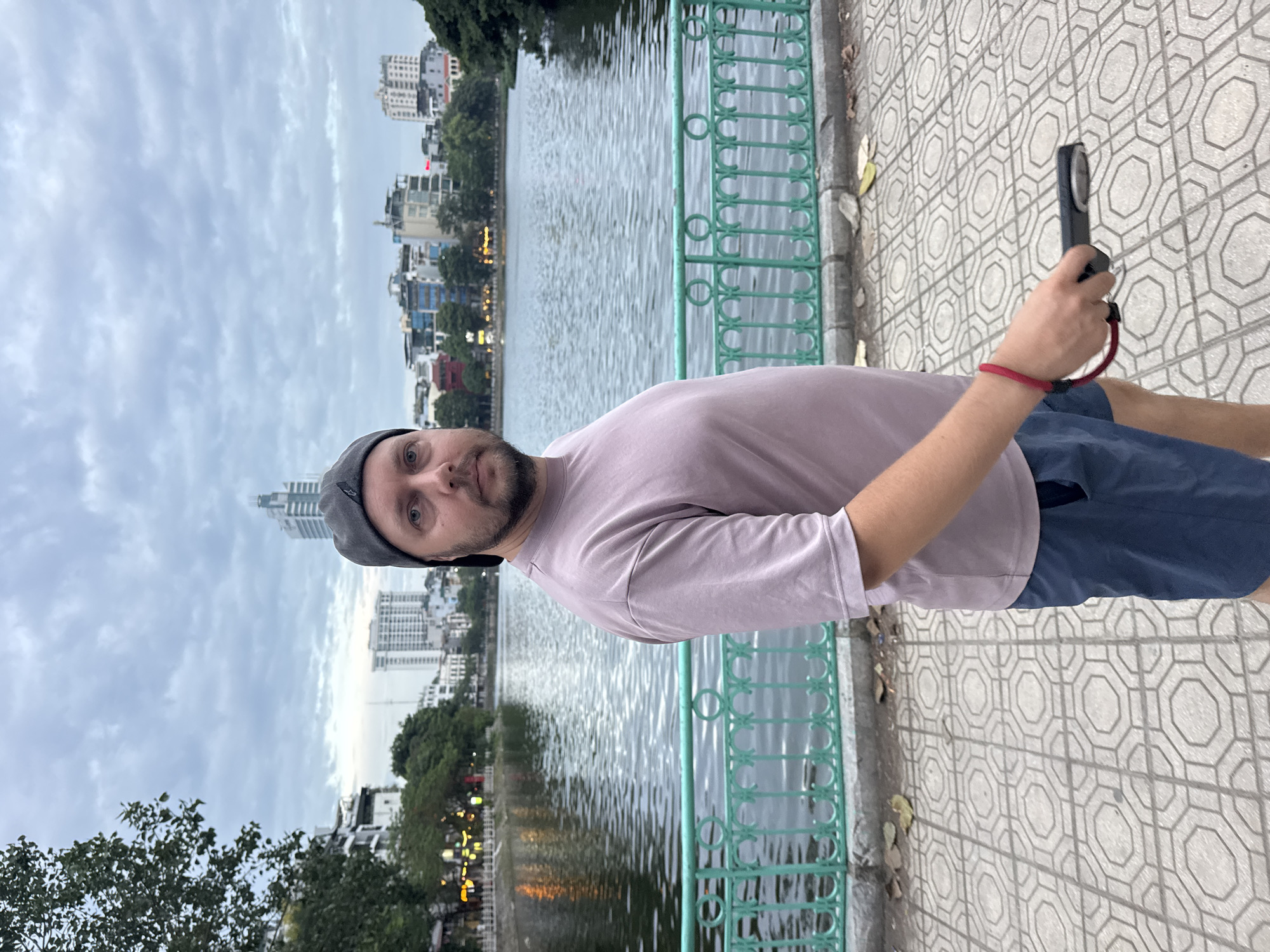 This media file captures a man standing near a body of water in what appears to be an urban setting. The man is wearing a light purple t-shirt, dark blue shorts, and a grey baseball cap. He is holding a dark-colored object, possibly a phone or camera, in his right hand. A red wristband is visible on his left wrist.
The background features a calm body of water reflecting the overcast sky. Across the water, a cityscape with various buildings, including some tall structures, is visible. Trees and streetlights can also be seen along the waterfront. A green metal railing runs along the edge of the paved area where the man is standing, separating him from the water. The paving stones have a repeating geometric pattern.
The sky is cloudy, suggesting it might be an overcast day or late afternoon. The lighting is soft, and there are reflections of lights in the water, hinting at the presence of artificial illumination even with the subdued daylight. The overall scene conveys a sense of urban exploration or a casual stroll in a city park or by a lake. The man's direct gaze towards the camera suggests he is aware of being photographed. The location context indicates this is Hà Nội, Vietnam, and the architecture and waterfront setting are consistent with such a location. No visible text is present in the image.