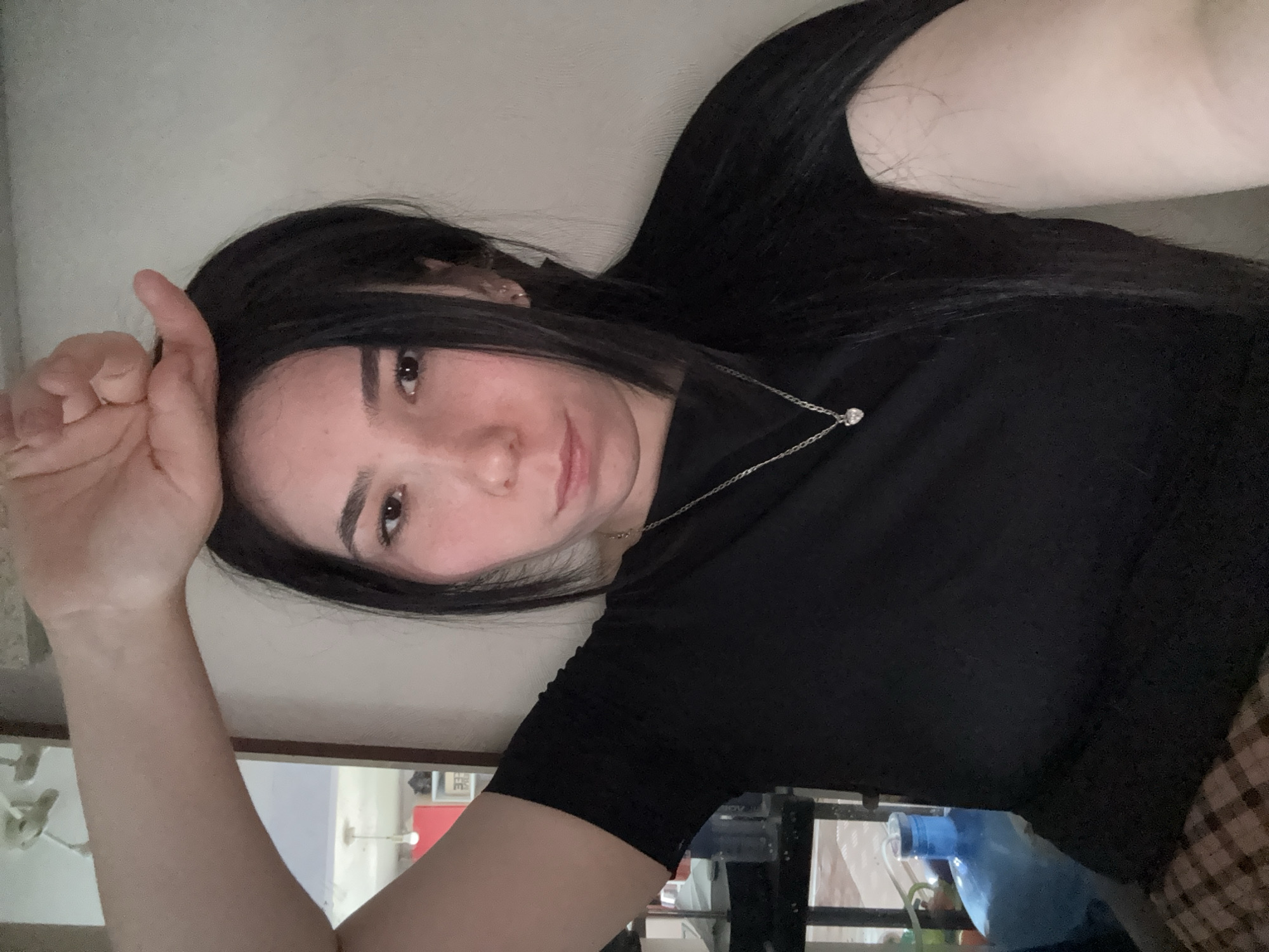 A young woman with long black hair and a silver necklace is posing for a selfie. She is wearing a black t-shirt and has her left arm raised with her hand near her forehead. The background is a plain wall. In the lower part of the image, there are objects like a blue bottle and a plaid pattern, suggesting an interior space. No information related to the city of Izhevsk, Russia, is identifiable in the image.
