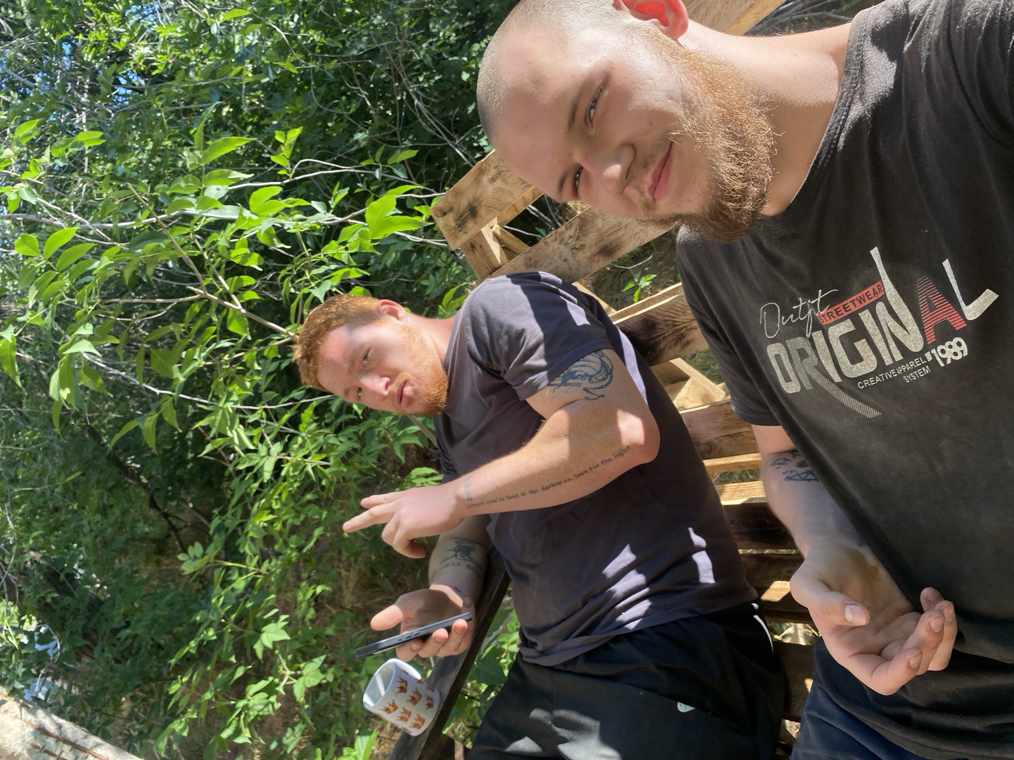 Two young men are seated on a wooden bench outdoors, surrounded by green foliage and trees. The man on the left, with red hair and a beard, is wearing a dark t-shirt and black shorts. He is holding a smartphone in his left hand and gesturing with his right hand. A mug with a crown pattern is visible to his left. The man on the right, with a shaved head and a beard, is wearing a dark t-shirt with a graphic print. He is looking towards the camera. The lighting suggests it is daytime.