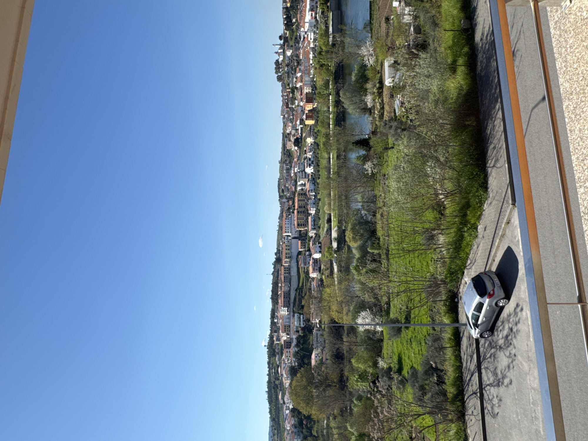 The image displays a panoramic view of Mirandela, Portugal, captured from an elevated position, likely a balcony, on a bright, clear day. The foreground features a metal railing, appearing to be stainless steel with a brown top bar, partially obscuring a textured, light-colored balcony floor. Immediately below, a paved area, consistent with a parking lot or street, contains a parked silver-grey hatchback car. Tree shadows are cast across this paved surface and the vehicle. A streetlight pole stands vertically adjacent to the paved area.
The midground reveals a lush green landscape with numerous trees and bushes, some exhibiting white blossoms, sloping down towards a calm river. The river reflects the clear blue sky. On the far bank of the river, additional green vegetation and what appear to be small agricultural plots with white-covered structures are visible.
The background presents the city of Mirandela, sprawling across undulating terrain. A dense arrangement of buildings with light-colored facades and red-tiled roofs dominates the hillside. A water tower is identifiable on a higher elevation to the left, and a prominent white structure with a red dome or roof stands on a hilltop to the right. The sky overhead is a uniform, clear blue with minimal cloud cover, indicating bright daylight. No people, specific actions, or interactions are discernible within the frame.