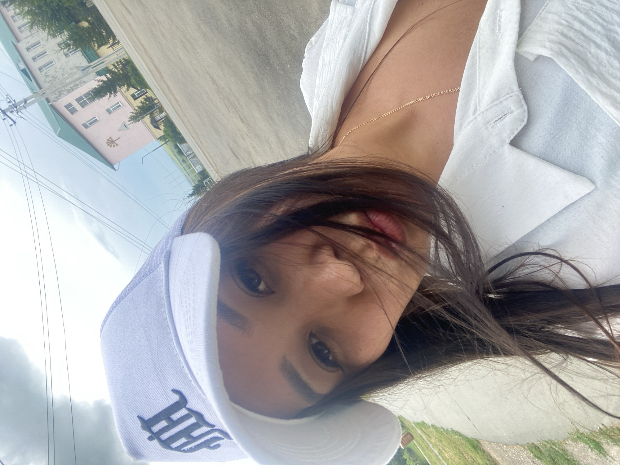 The image is a close-up selfie of a woman wearing a white baseball cap with a dark logo and a white collared shirt. She has long, dark hair and is looking at the camera. Behind her, there is a road, some grass, and buildings. The sky is cloudy. The setting appears to be an outdoor urban or suburban environment.