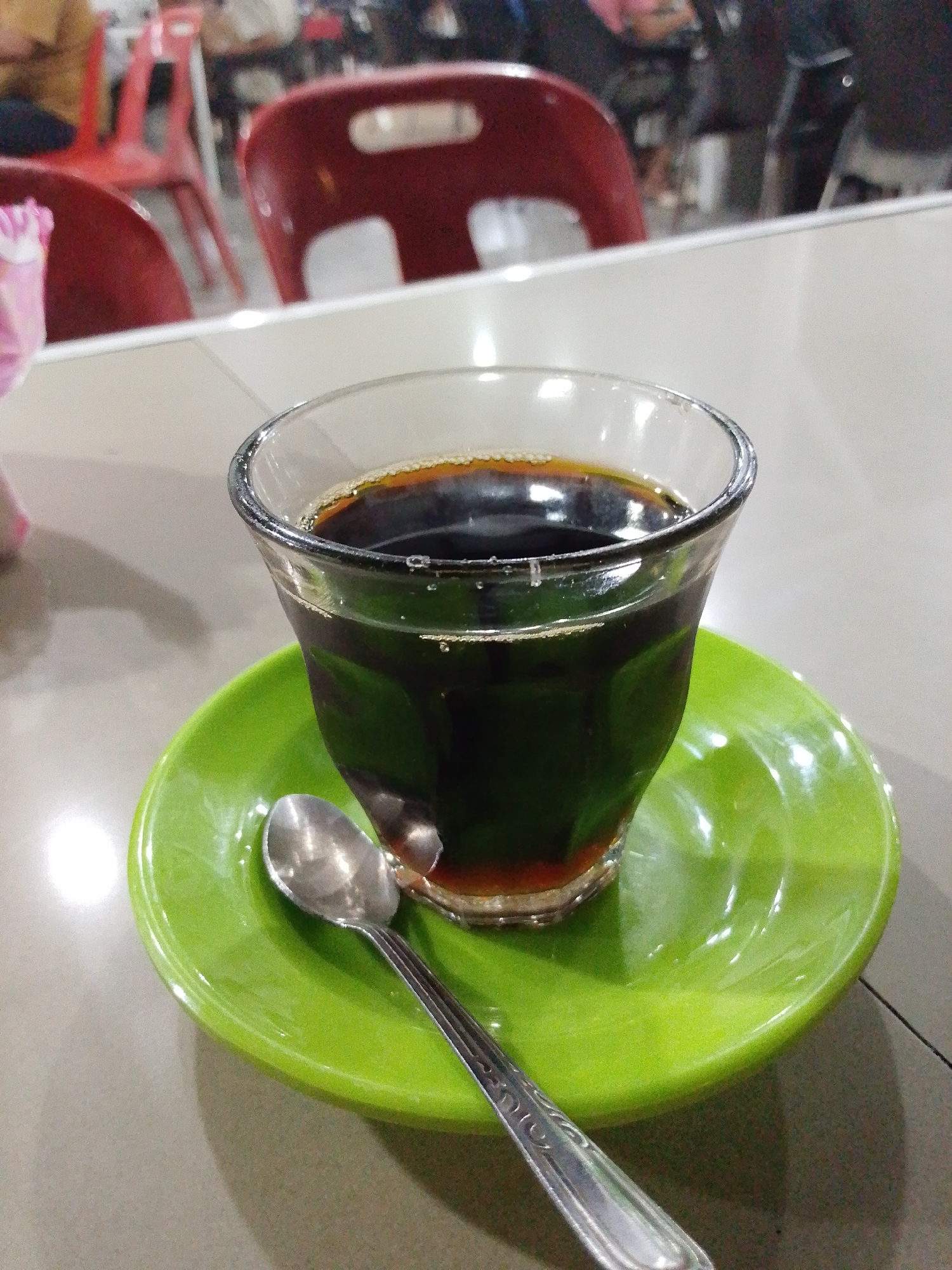 The image captures a glass of dark liquid, likely coffee, placed on a bright green saucer alongside a small silver spoon. The setting appears to be a table at a restaurant or cafe, indicated by the presence of chairs and indistinct figures in the background. The location is Lhokseumawe, Indonesia, which is mentioned. The foreground shows a clear glass filled with a dark fluid, probably coffee, resting on a vivid green saucer with a spoon next to it. The tabletop is light-colored, reflecting light. The background features red and black chairs and blurred figures suggesting an occupied dining area in Lhokseumawe.