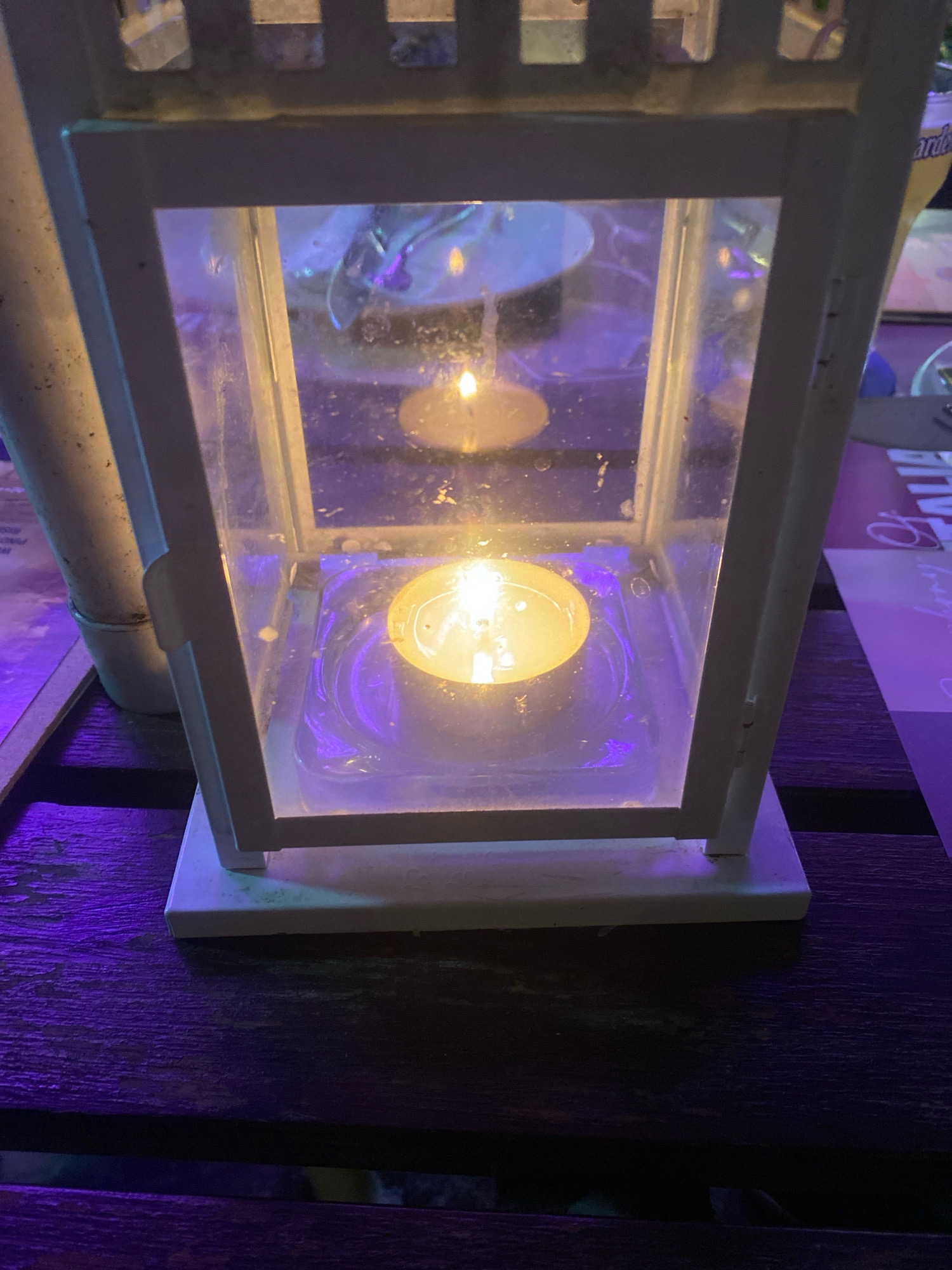 This is a close-up, vertical, eye-level photograph of a white lantern with a lit candle inside. The lantern has a white frame and clear glass panels. The candle, a small tea light, is illuminated and centered within the lower section of the lantern. The flame is bright yellow and casts a warm glow. The lantern sits on a dark, wooden surface, possibly a table, which has a purple ambient light reflecting onto it. In the background, through the glass panels of the lantern, there are blurry reflections of lights and what appear to be other objects, suggesting an indoor or outdoor setting at night. The overall impression is one of a dimly lit, atmospheric scene. The photograph was taken in Kuala Lumpur, Malaysia.