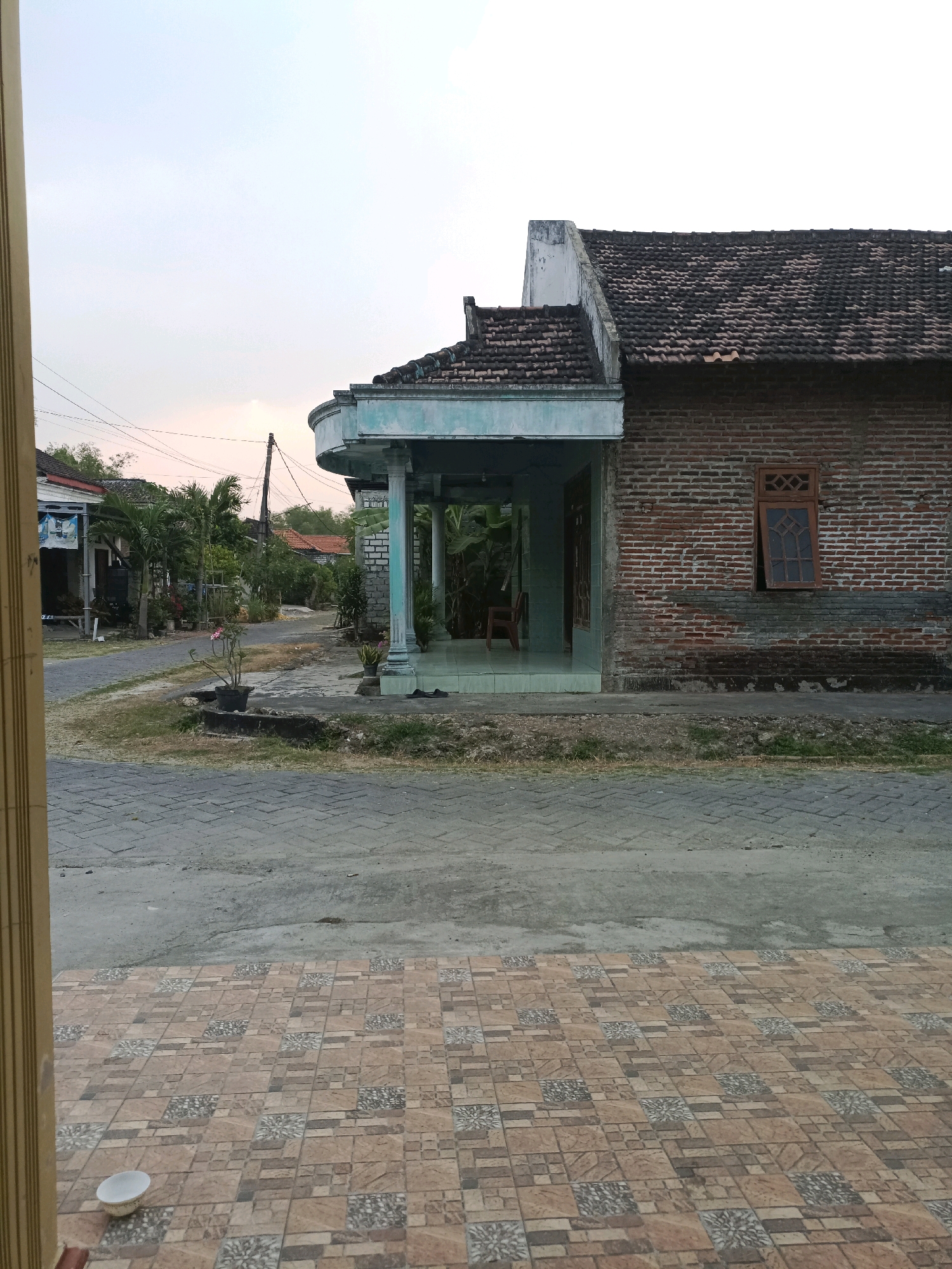 The image depicts a residential street scene in Rancang Kencono, Indonesia, with a focus on the exterior of a brick house. The house has a tiled roof, a covered porch with pillars, and a wooden window with a decorative lattice pattern. Adjacent to the house is a paved road and sidewalk. Various potted plants and trees are visible throughout the scene. The sky is overcast, suggesting it may be dawn or dusk. There are no people present in the image.