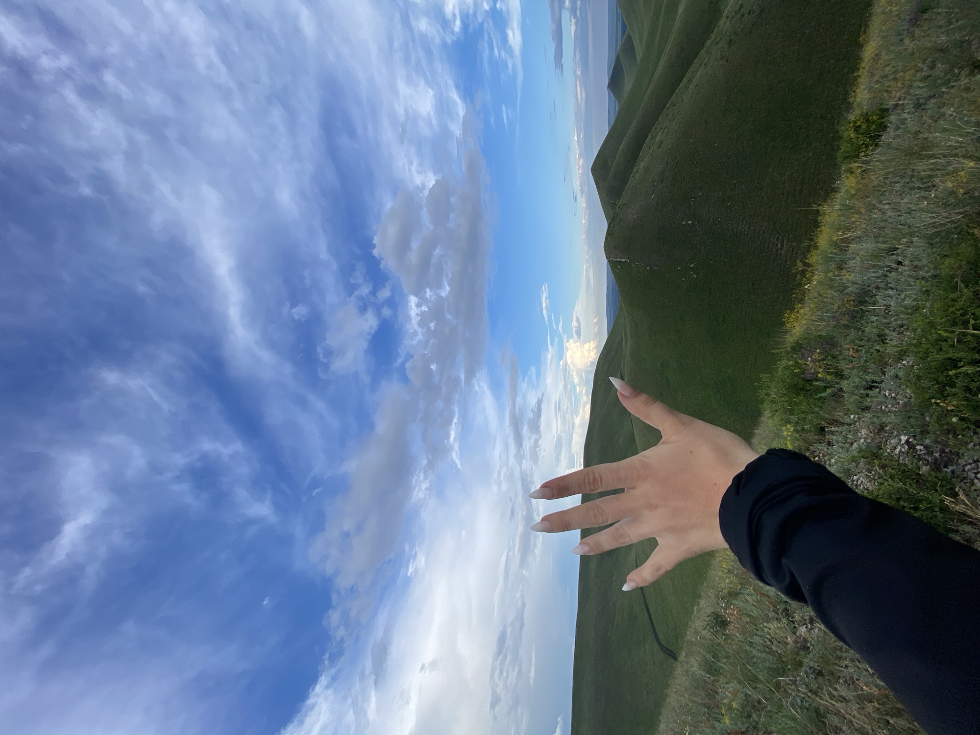 The image depicts a landscape of rolling green hills under a blue sky with scattered white clouds. In the foreground, a person's left hand, adorned with long, white, pointed fingernails, is raised, with fingers spread open. The person is wearing a black long-sleeved garment. The terrain is covered in lush green grass, with some smaller plants and wildflowers visible on the slopes. A faint track is visible winding up one of the hills in the mid-ground. The horizon line shows a hazy distant view of more hills and possibly water. The overall impression is of a natural, outdoor setting, likely in the countryside surrounding Verkhneozernoe, Russia.
