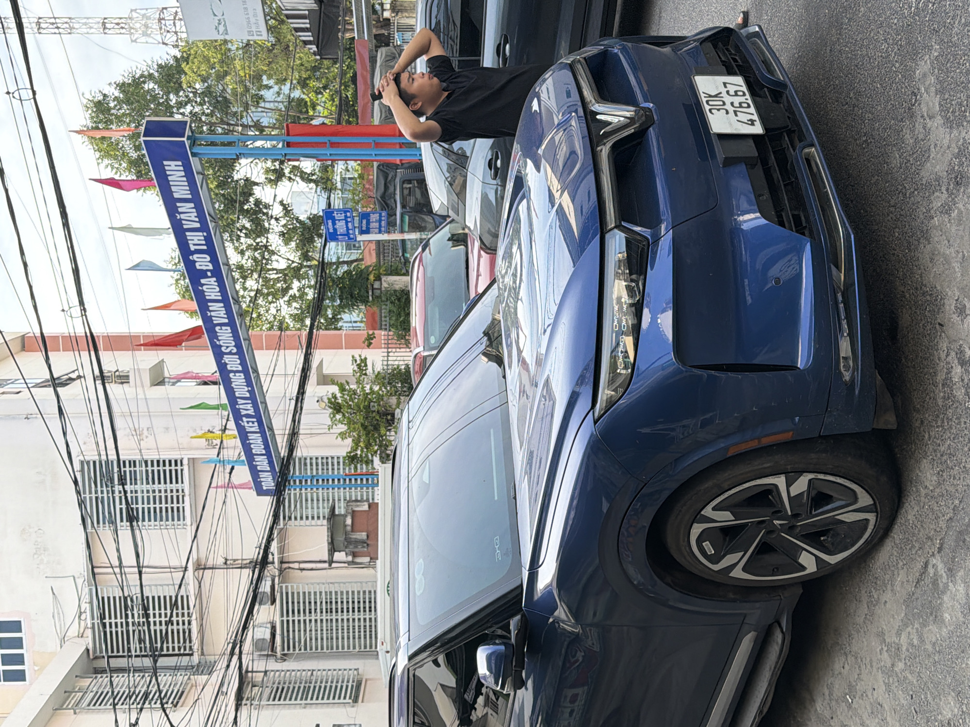 In Nha Trang, Vietnam, a blue electric vehicle is parked on a street. The car, a VinFast VF e34, has a license plate that reads "30K 476.67". A young man is standing to the right of the car, with his hands on his head, looking away from the camera. In the background, there is a building with shutters on the windows, a blue sign with Vietnamese text, and overhead electrical wires. Other cars are also visible, including a red SUV and a gray sedan. The overall scene depicts a typical street view in a Vietnamese city.
