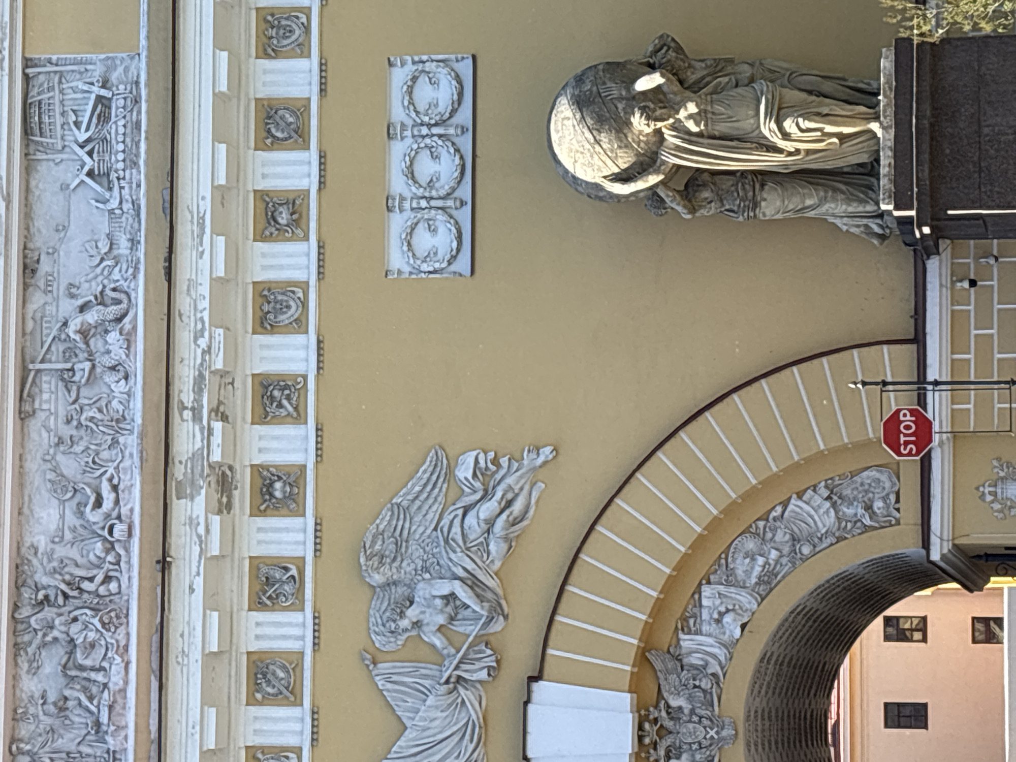 This image shows a close-up of the facade of the General Staff Building in Palace Square, Saint Petersburg, Russia. The building's yellow stucco walls are adorned with elaborate bas-relief sculptures and architectural details.
In the upper section, a large, intricate frieze depicts a mythological or historical scene, featuring numerous figures and naval elements, including a ship and an anchor. Below this, a band of decorative panels showcases various heraldic symbols and emblems, such as shields, anchors, and crossed weapons.
To the left, a winged figure, possibly an angel or allegory, is depicted in motion, holding a banner or fabric. To the right of this figure, a rectangular plaque features three circular wreaths.
Dominating the lower right portion of the image is a monumental sculpture of several female figures supporting a large globe, likely an allegory for Atlas. These figures are draped in classical robes.
An arched entryway is visible in the lower left, with further sculpted decorations along its curve and within its recess. A stop sign is prominently placed on a pole to the right of the archway. In the background, through the archway, glimpses of a pink building with windows can be seen. The overall architectural style suggests Neoclassical influences.