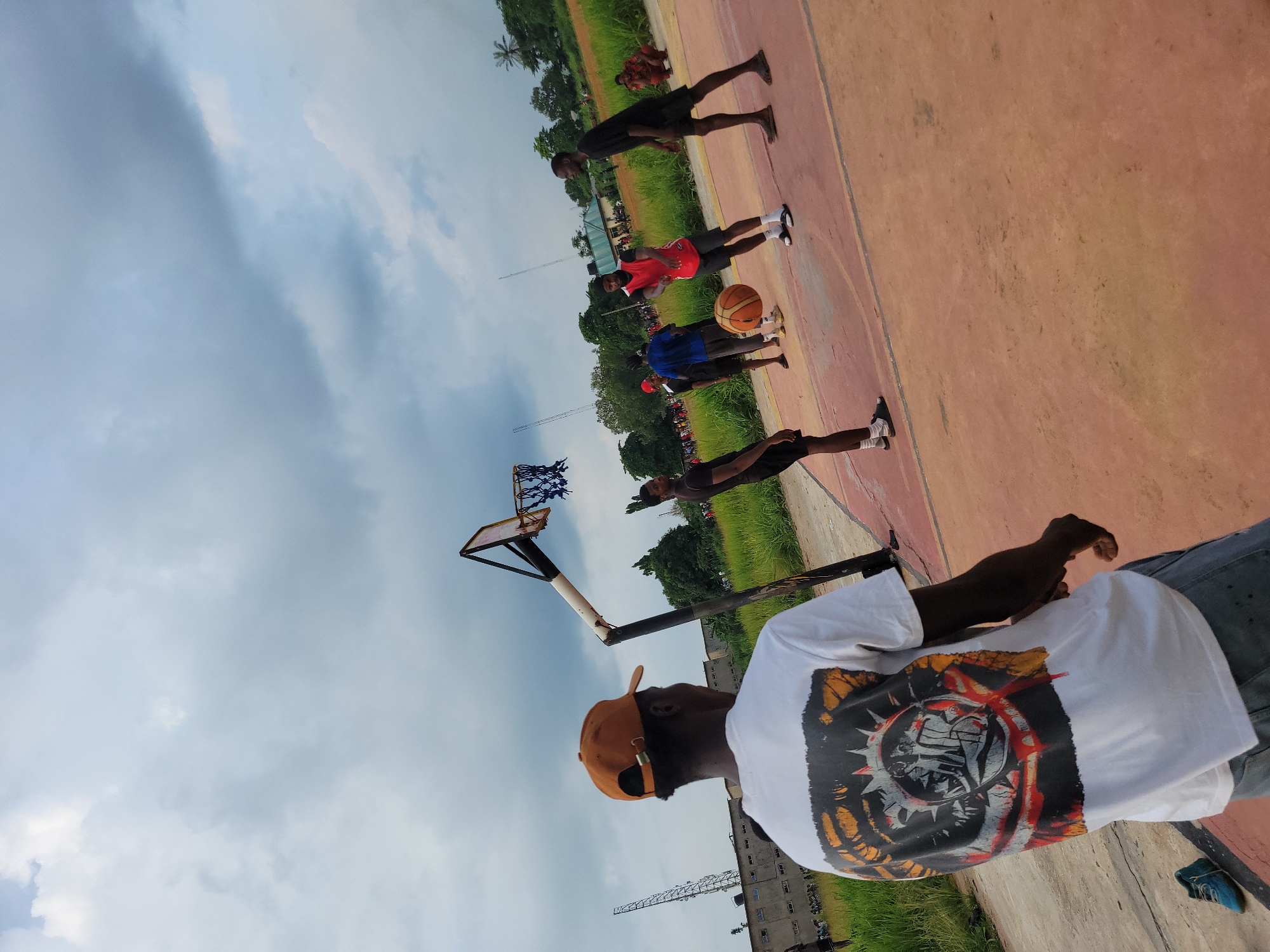 This image captures an outdoor basketball court scene in Egbagu, Nigeria, under a heavily clouded, grey sky. In the foreground, facing away from the viewer, a dark-skinned male wears a brown baseball cap and a white t-shirt featuring a large, multi-colored graphic design on the back, depicting an intricate, spiky circular motif with abstract figures. He wears grey lower garments, and a single blue shoe is visible on the dirt ground near his left foot.
On the reddish-orange asphalt basketball court, which shows signs of wear and faded lines, multiple dark-skinned males are engaged in a game. Centrally positioned, a male in a red sleeveless jersey and black shorts actively dribbles an orange-brown basketball with his right hand, wearing white socks and white slides. To his immediate right, another male in a dark t-shirt and dark shorts stands, observing the play. To the left of the dribbling player, a male in a blue t-shirt and grey pants is partially visible. Further to the left, another male in a dark t-shirt and shorts stands near the court's edge. On the far right, a male in a black t-shirt and black shorts stands, observing the action, appearing barefoot or in very thin sandals.
A single, weathered basketball hoop with a white backboard, a black support pole, and a blue rope net is positioned on the left side of the court. Surrounding the court is abundant green grass and various trees. In the background on the far left, a tall metal lattice tower is visible next to a block of grey buildings. A smaller building with a dark roof is centrally located in the mid-distance. On the far right background, a person in an orange-brown garment is crouched on the ground, observing the scene.