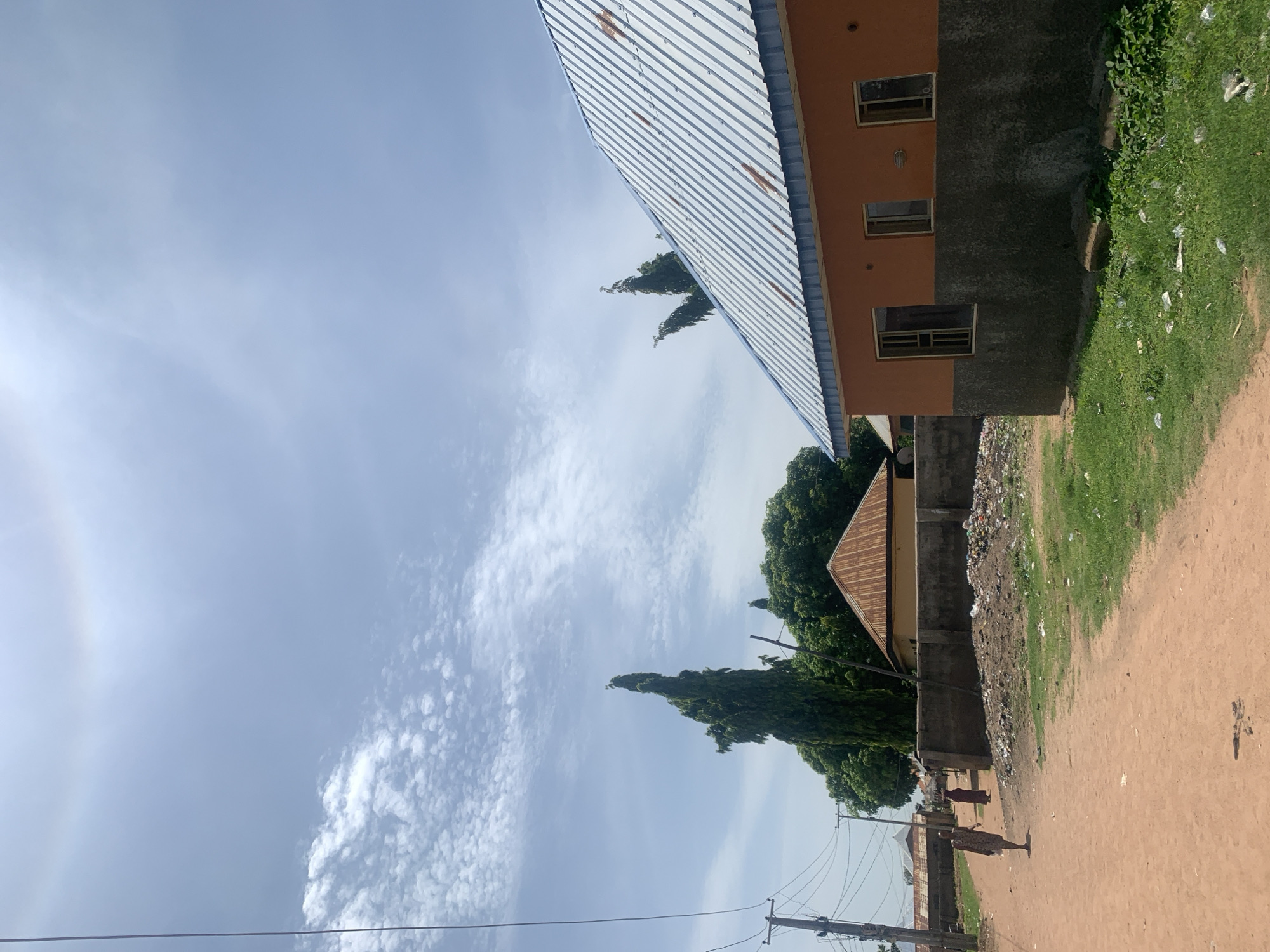 In Jauro Yinu, Nigeria, a dirt road curves through a settlement under a partly cloudy sky. To the right, an orange building with a corrugated metal roof features three windows. The building's lower half is dark gray. A pile of garbage is visible on the ground to the left of the building. Further down the road, a tall, slender green tree stands prominently, dwarfing other trees and buildings. Two women are walking on the road, one in a patterned dress and the other in a maroon garment. Utility poles with power lines are present along the left side of the road. The environment appears dry, with patches of green grass and scattered debris.