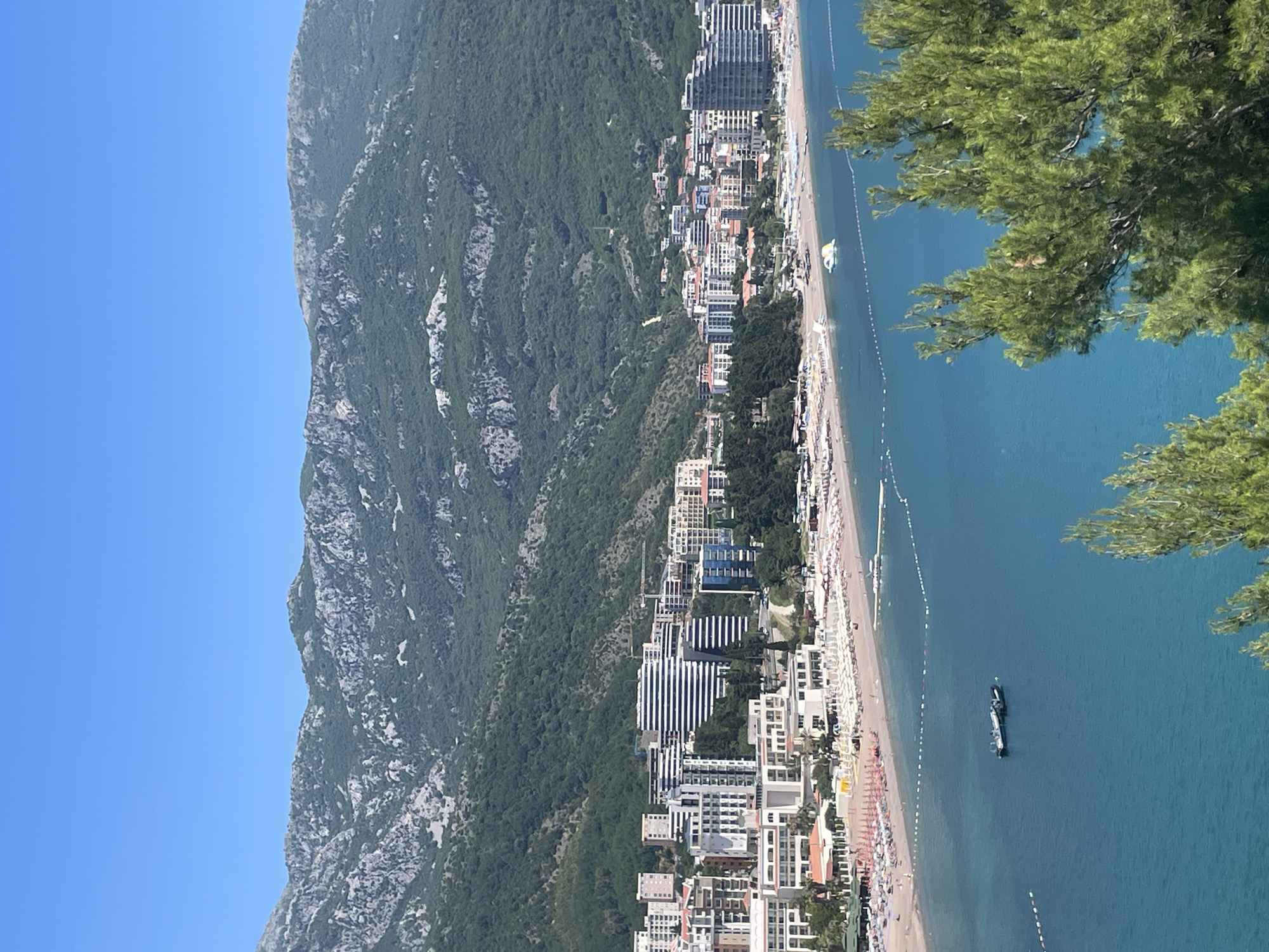 An eye-level, vertical shot reveals a mountainous landscape merging into a coastal city. To the left, a mountain covered with vegetation rises against a clear blue sky. As the mountain descends, it meets a developed coastline, presumably in the city of Boreti, Montenegro. Buildings of varying heights and architectural styles line the coast, juxtaposed with greenery. A sandy beach, densely populated with beachgoers, stretches along the waterfront. The blue sea extends from the beach, dotted with a single boat and marked by what appear to be safety buoys. The right side of the image features a green coniferous tree.